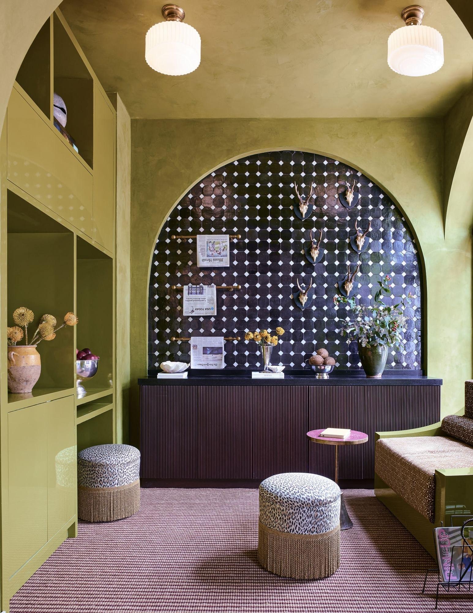 A hotel lounge corner with olive green walls, a dark tiled archway hung with faux antler trophies, leopard print stools, and a magazine rack.