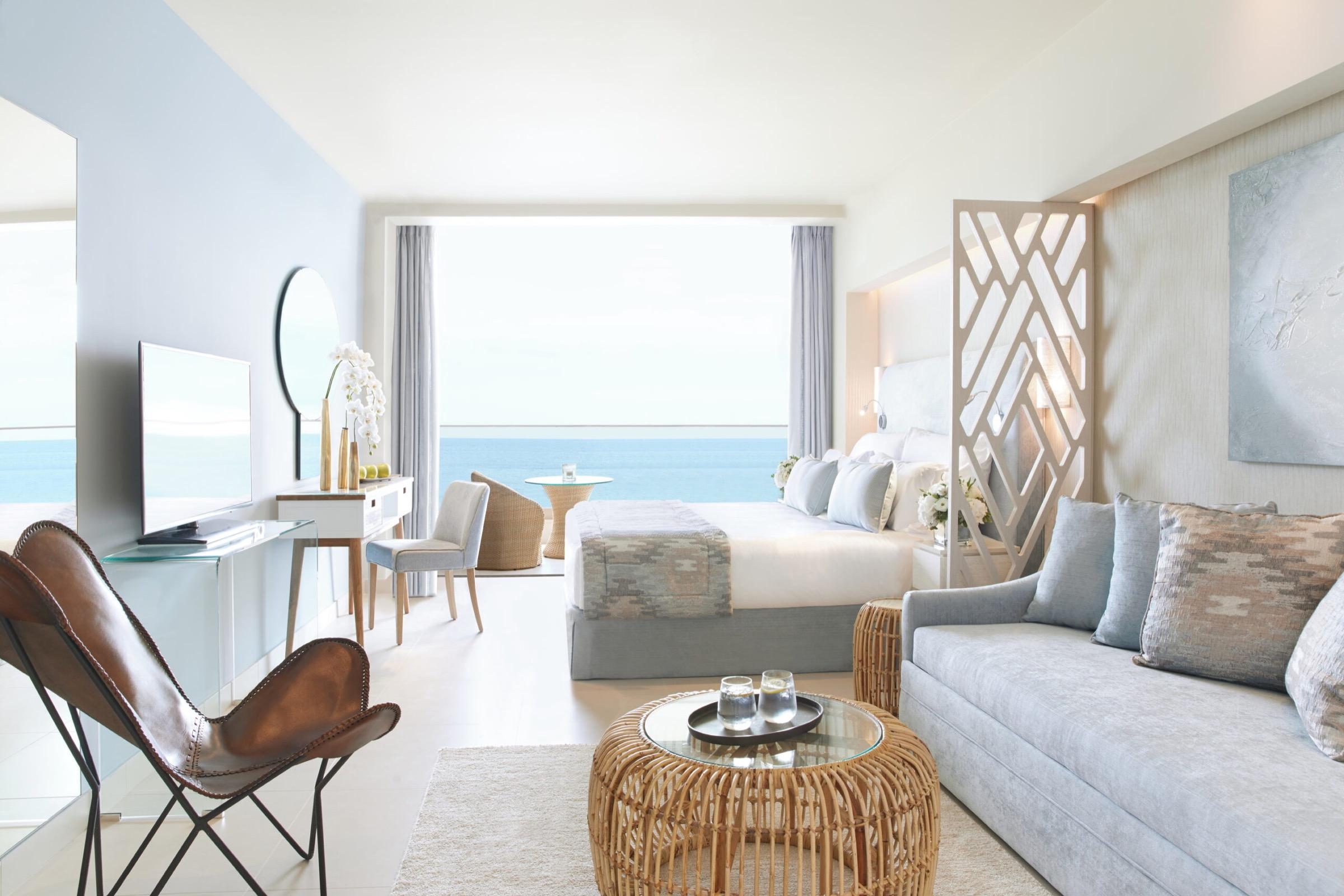 A hotel suite with a bed, sofa and television facing a glass door open to a sea view, with a round mirror and dressing table to one side.