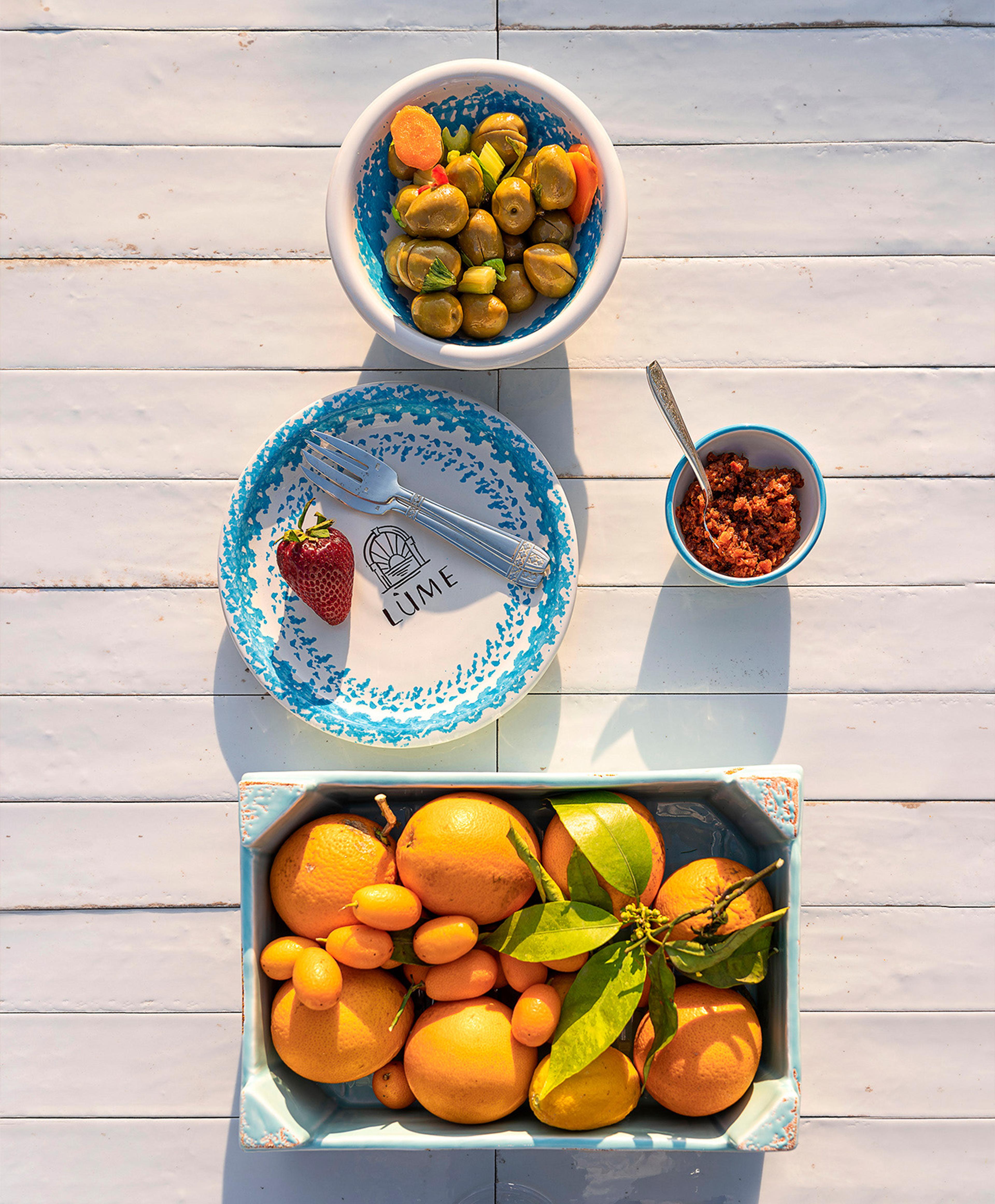A bowl of olives and carrots, a small bowl of nduja, a plate with a strawberry and fork, and a crate of oranges and kumquats, arranged on a white wooden table and shot from above.