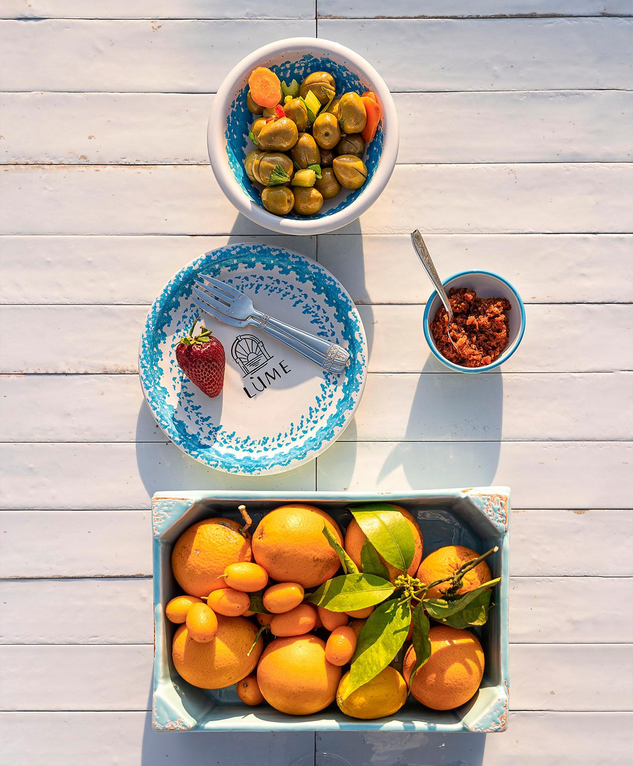 A bowl of olives and carrots, a small bowl of nduja, a plate with a strawberry and fork, and a crate of oranges and kumquats, arranged on a white wooden table and shot from above.