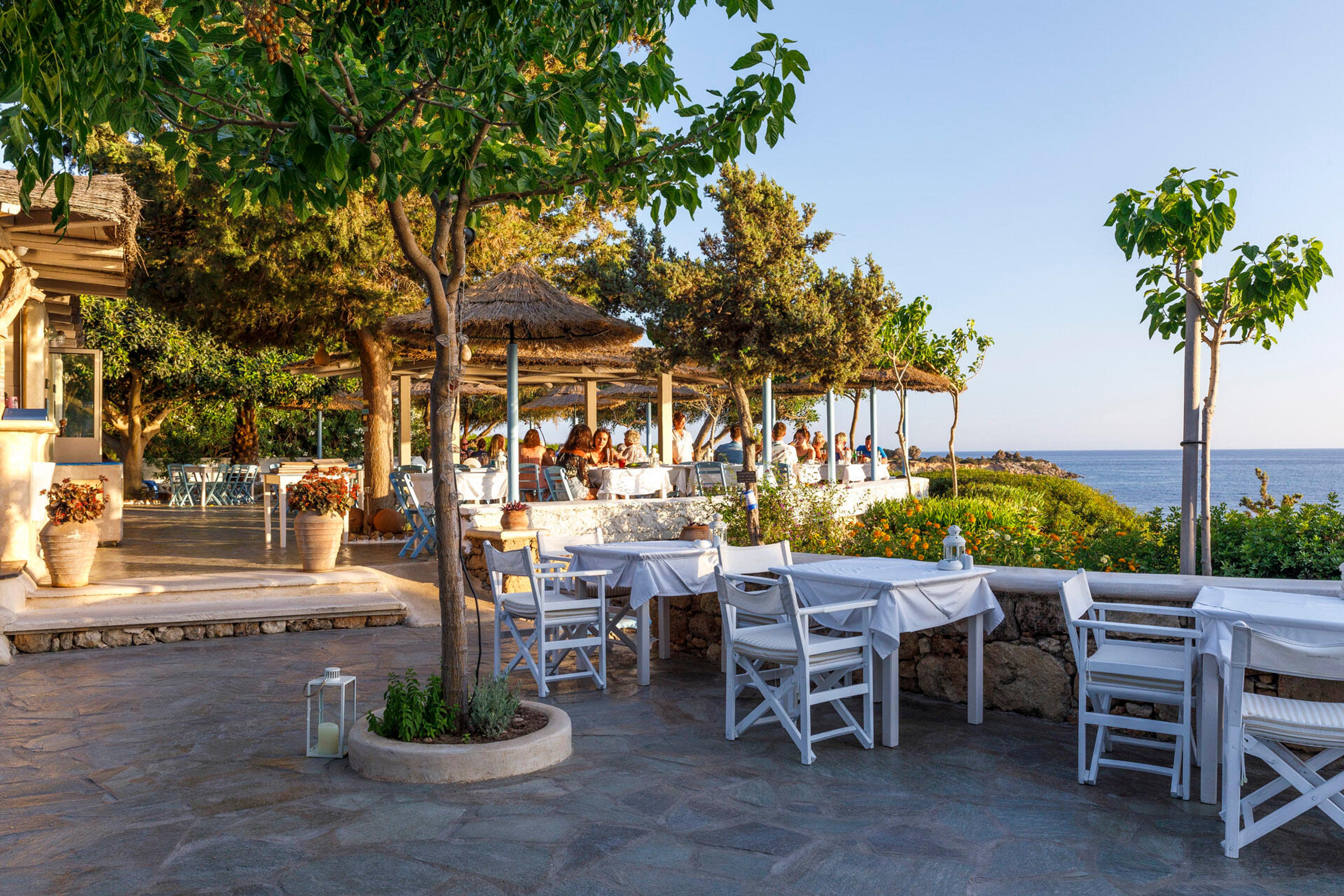 White tables and chairs on a stone garden terrace under trees and thatched shades, with the sea beyond