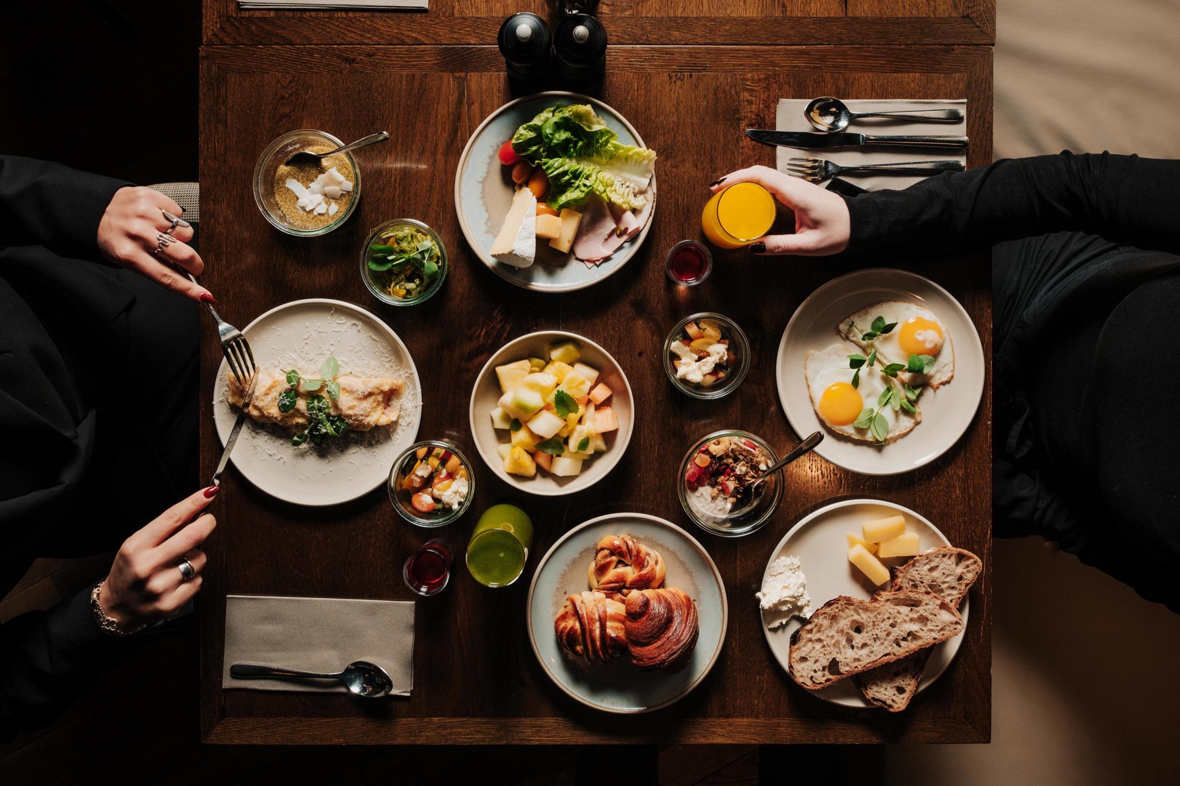 An overhead view of a wooden table set with plates of eggs, pastries, bread, cheese and fruit, with two people's hands and arms reaching in from either side.