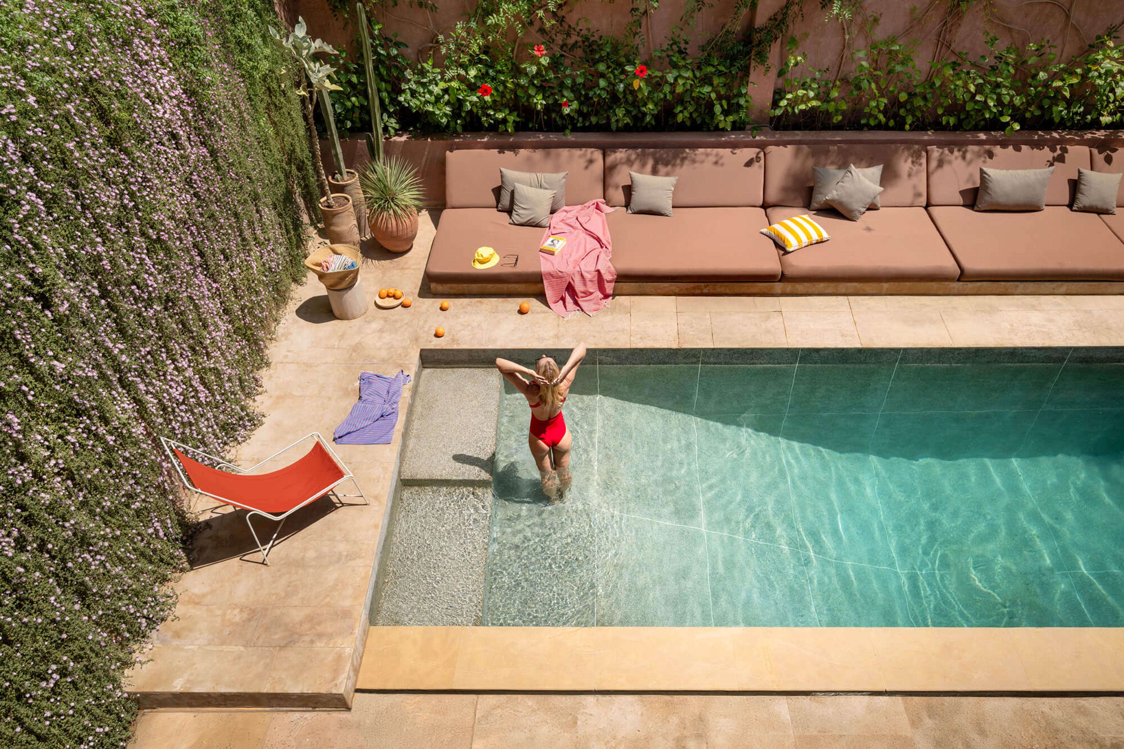 a woman in a red swimsuit, seen from behind, stepping down into a swimming pool beside a row of cushioned loungers and a wall of small purple flowers