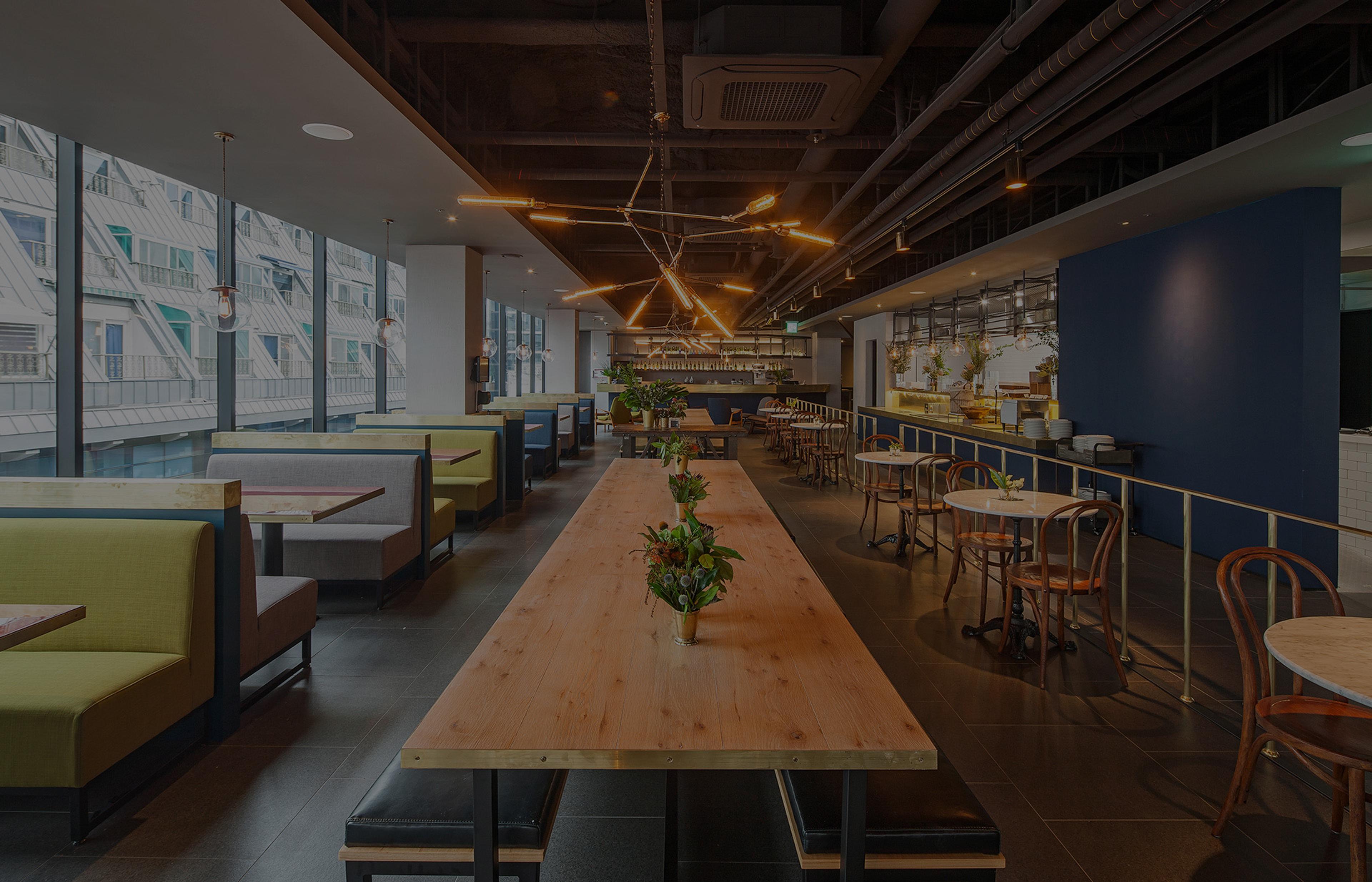 A long wooden communal table set with a small flower arrangement, running through a bright dining room with booth seating, pendant lights and windows looking onto a building across the street.