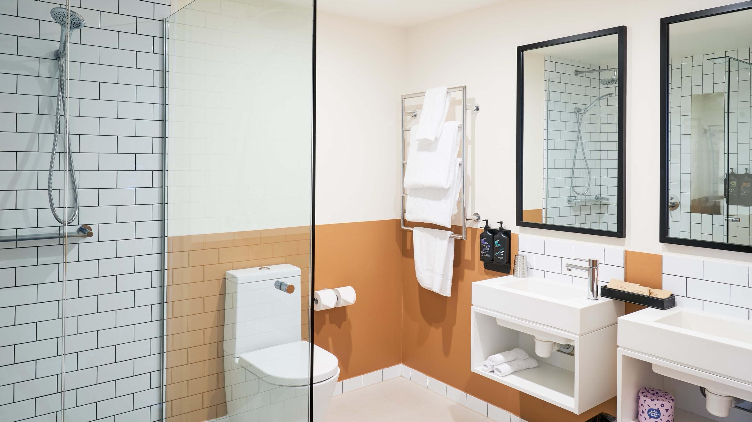 A bathroom with a glass shower, a toilet, a heated towel rail and a double vanity with two mirrors against a burnt orange wall.