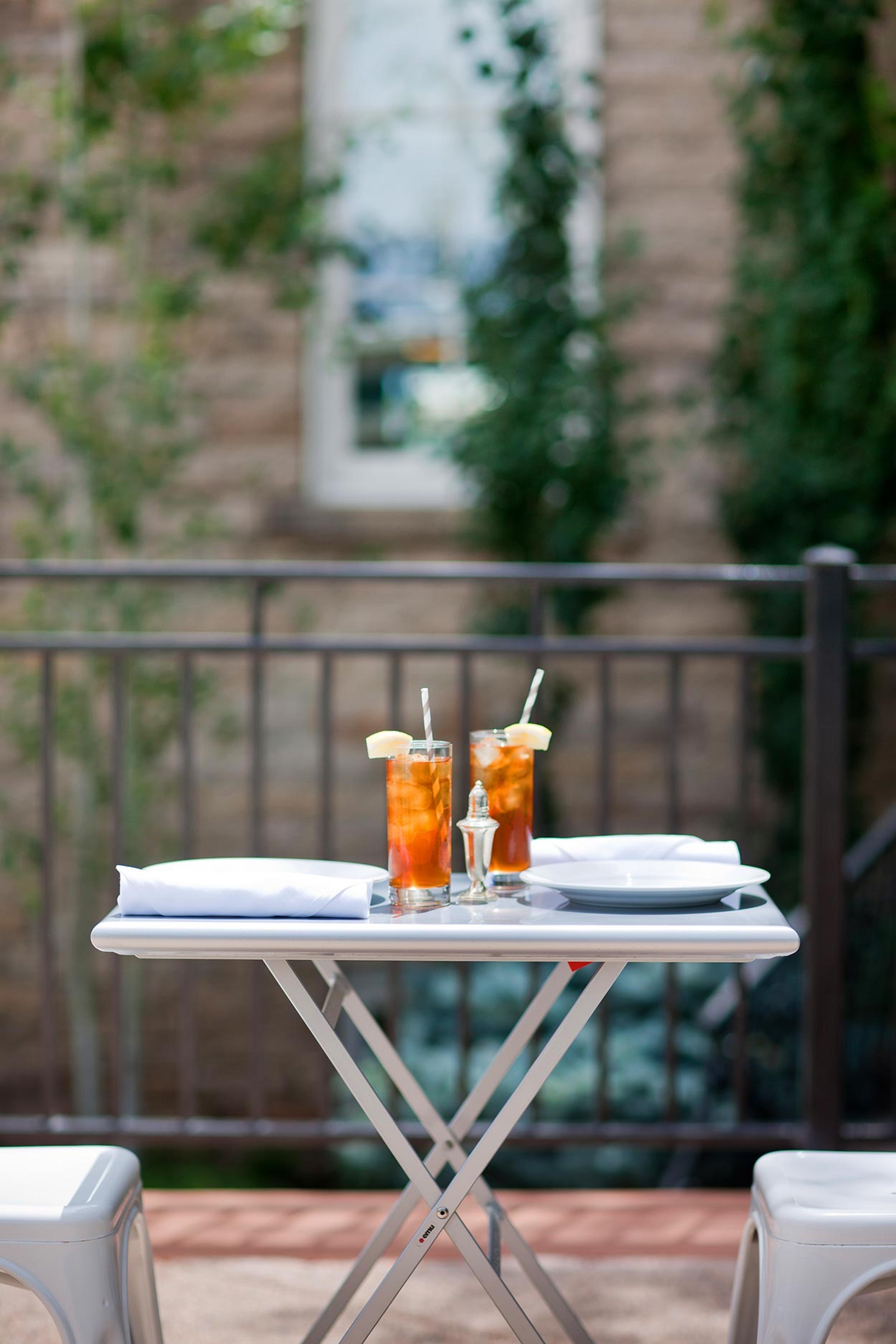 A small outdoor table set with two iced teas, a folded napkin and a plate, beside a wrought iron railing with greenery and the building's facade behind.