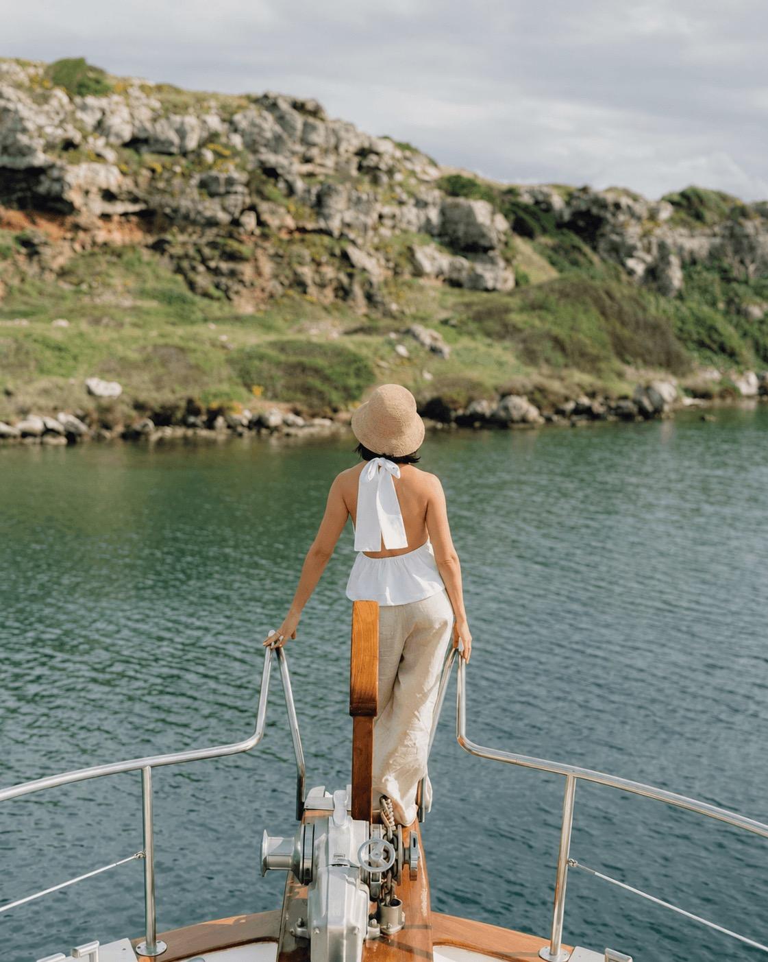A woman in a sun hat stands at the bow of a boat, looking out over the water to a rocky, scrub-covered coastline.