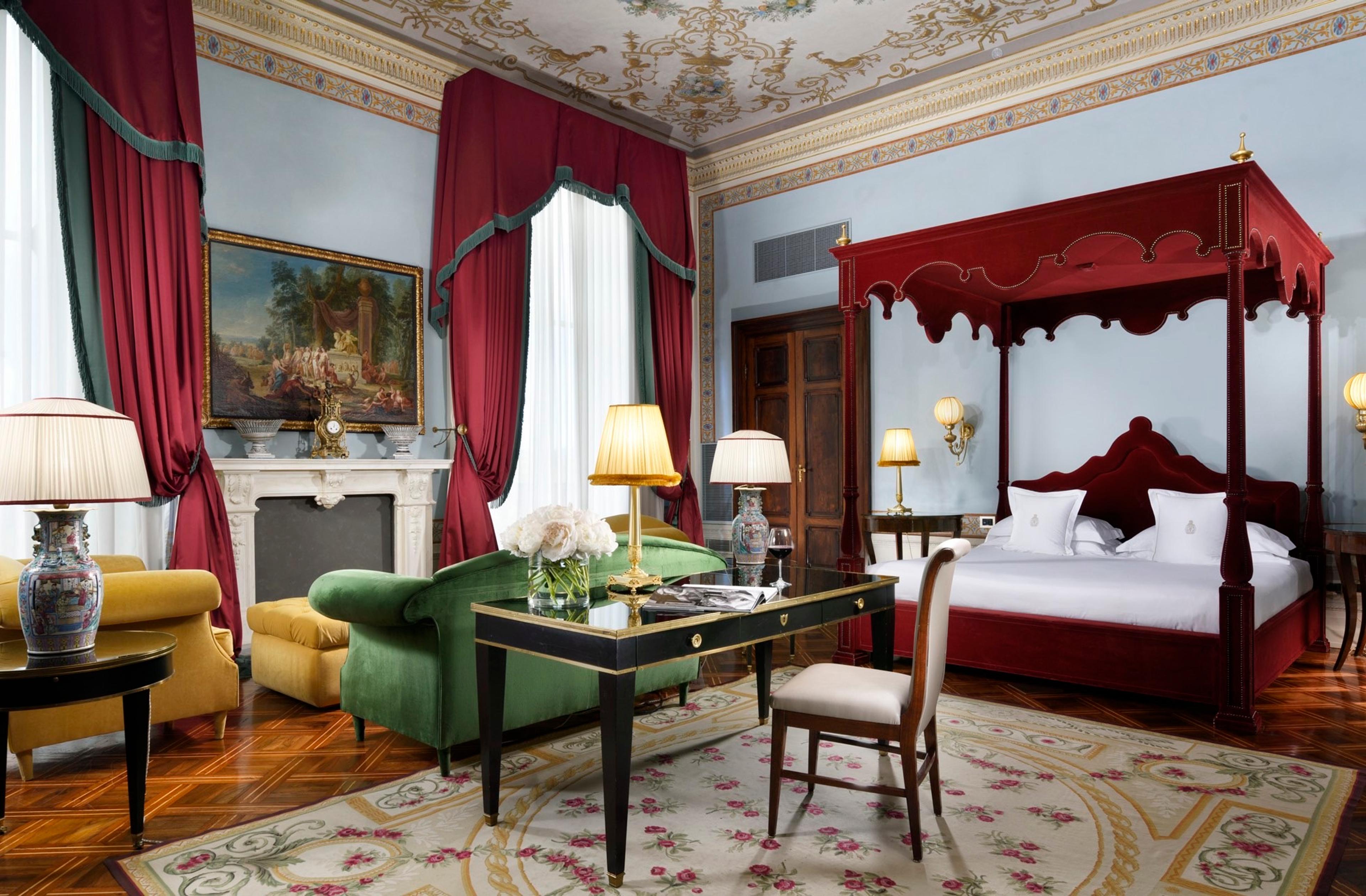 A large bedroom with a red four-poster bed, a painted ceiling, a green velvet sofa, a yellow armchair and a writing desk on a floral rug.
