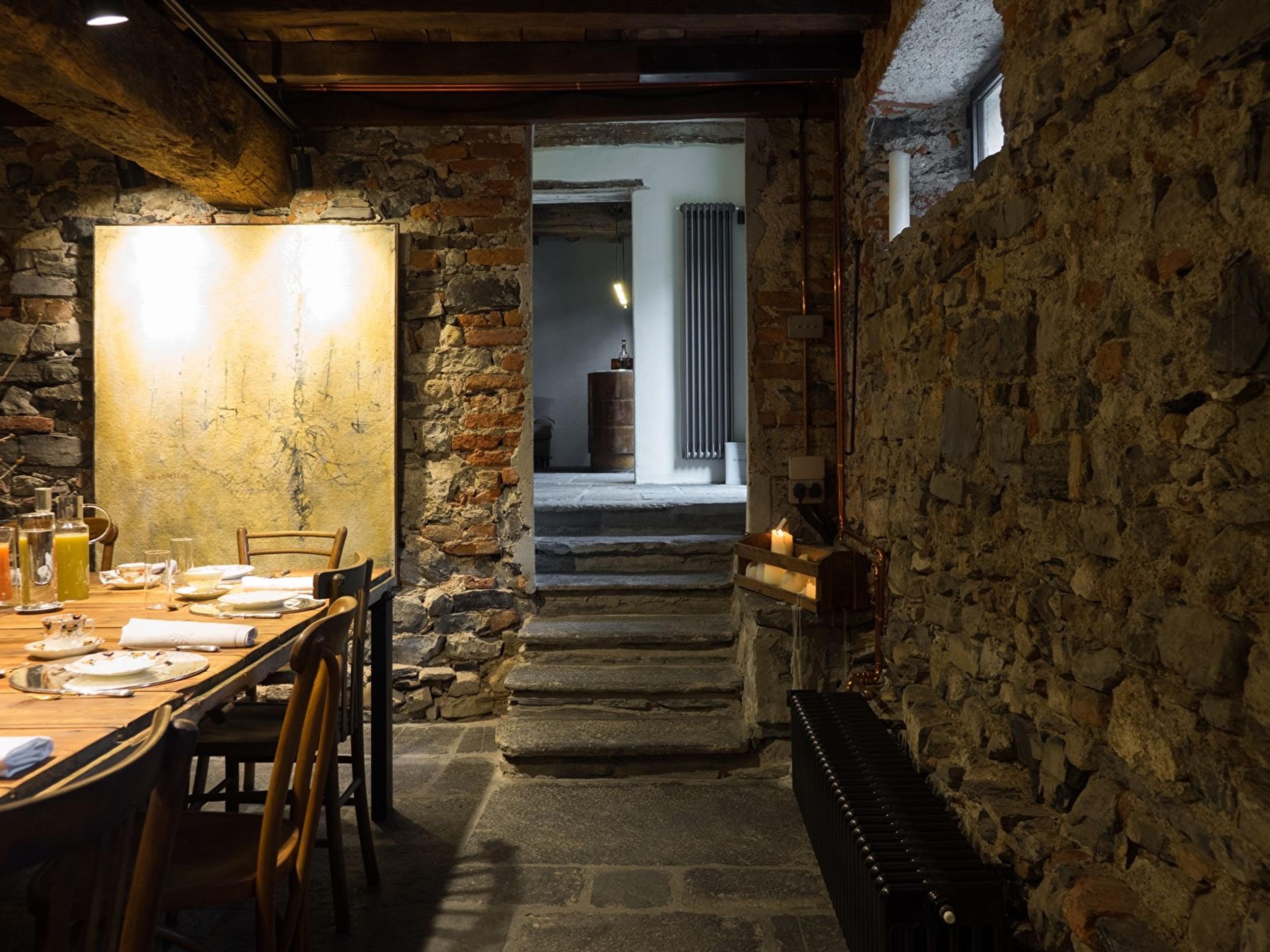 a stone-walled dining room with a set table, steps leading up to a lit room beyond, and a candle burning in an alcove