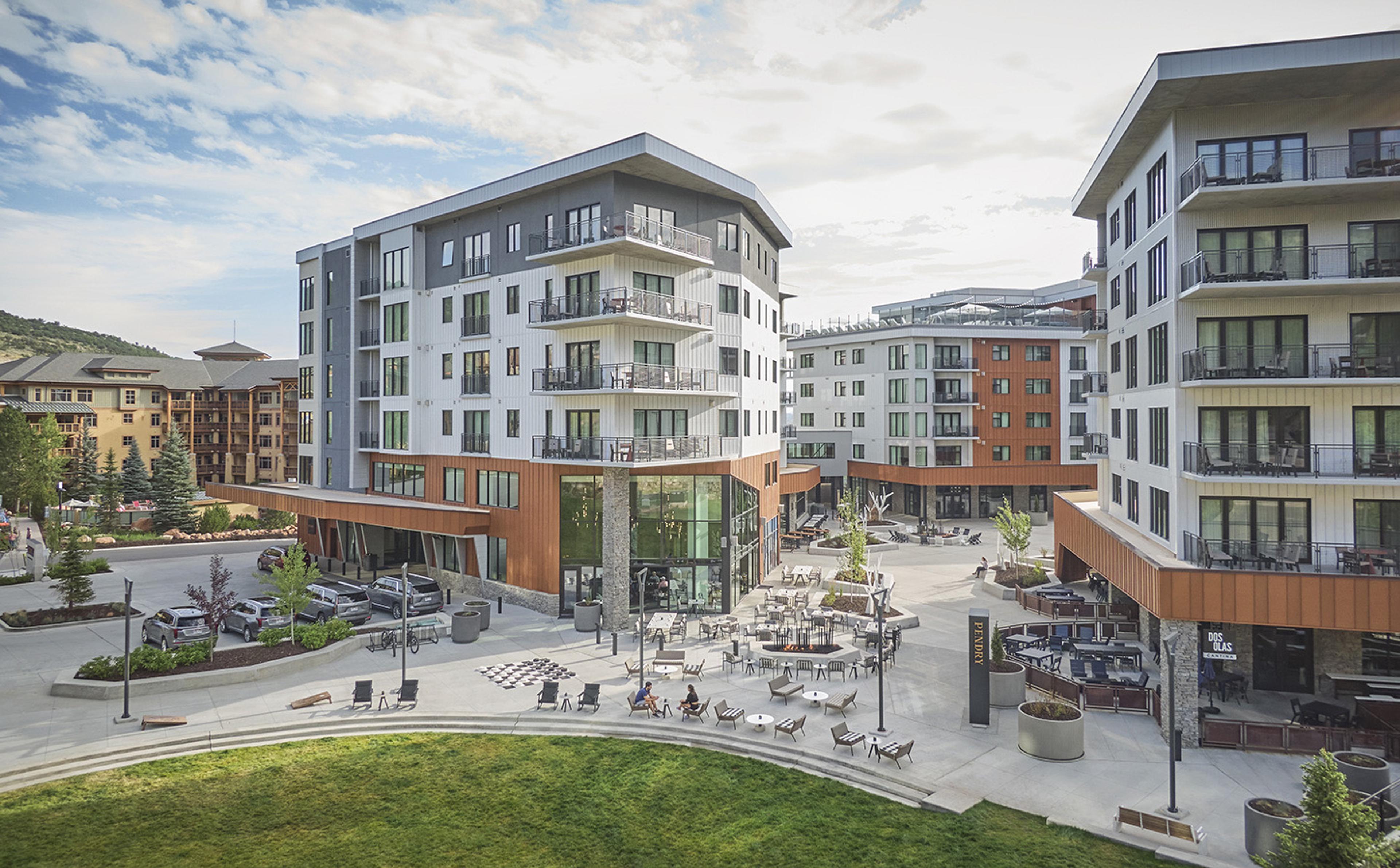 Aerial view of two multi-storey buildings with white panels and orange wood cladding, around a paved courtyard with tables, chairs and a few people, and a strip of lawn in the foreground.