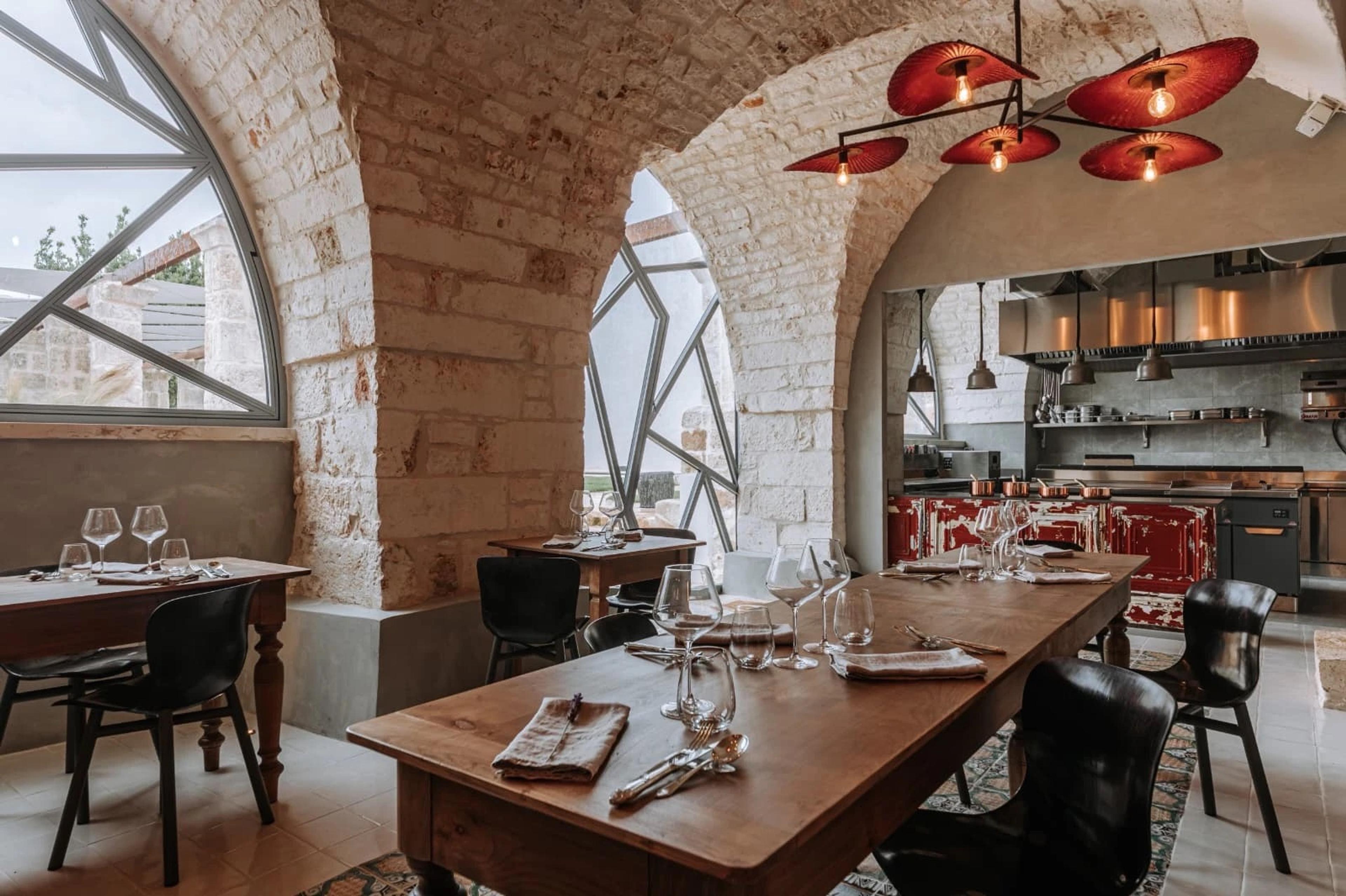 A vaulted stone dining room with wooden tables set with wine glasses and folded napkins, red pendant lamps overhead, and an open kitchen beyond.