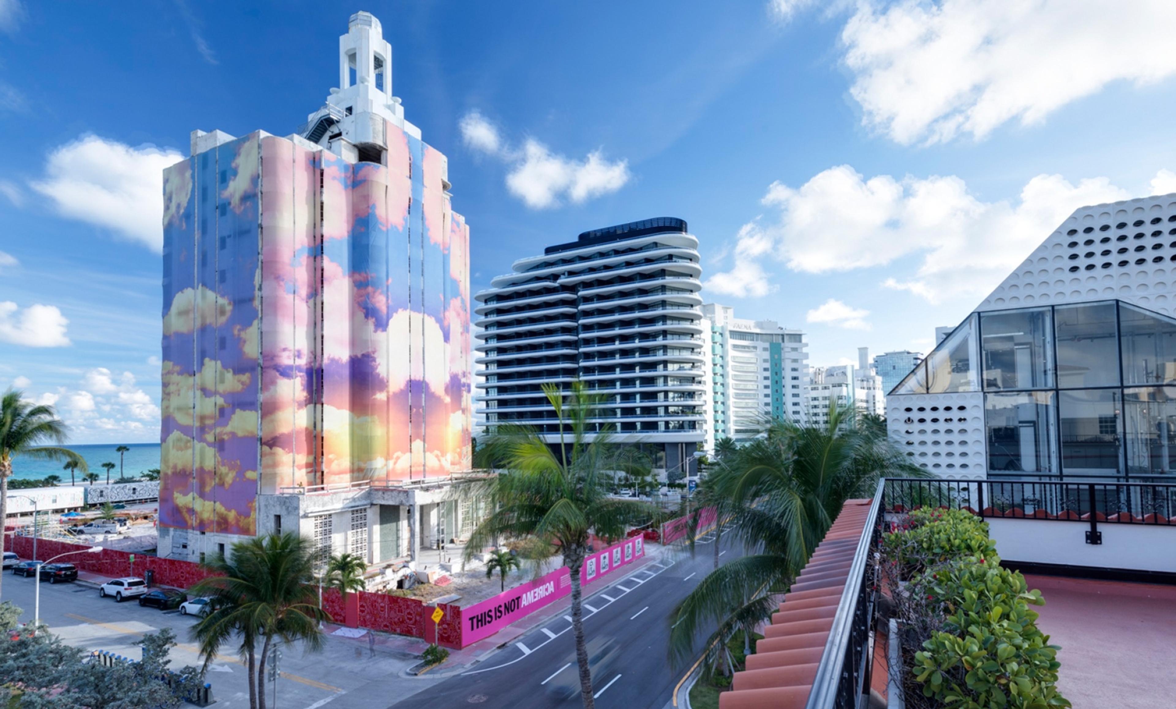 A white-topped tower on a street, its upper floors wrapped in a painted sky of pink and blue clouds, with palm trees, a newer tower behind it and a pink hoarding at its base.