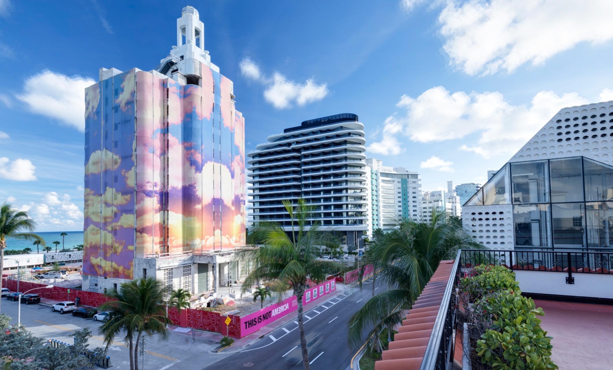 A white-topped tower on a street, its upper floors wrapped in a painted sky of pink and blue clouds, with palm trees, a newer tower behind it and a pink hoarding at its base.