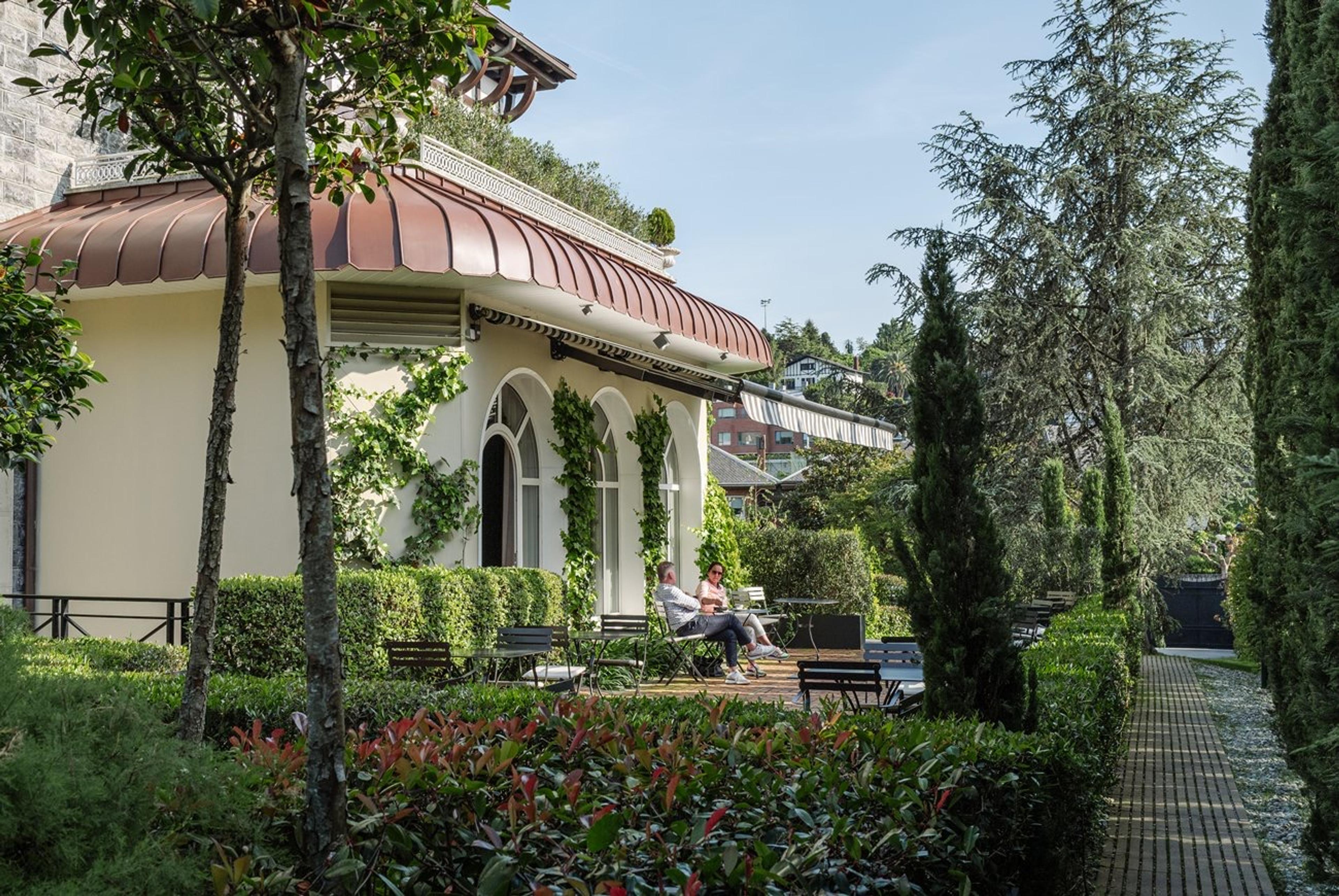 A domed terrace with arched windows, outdoor tables and chairs, and two people seated at a distance among trees.