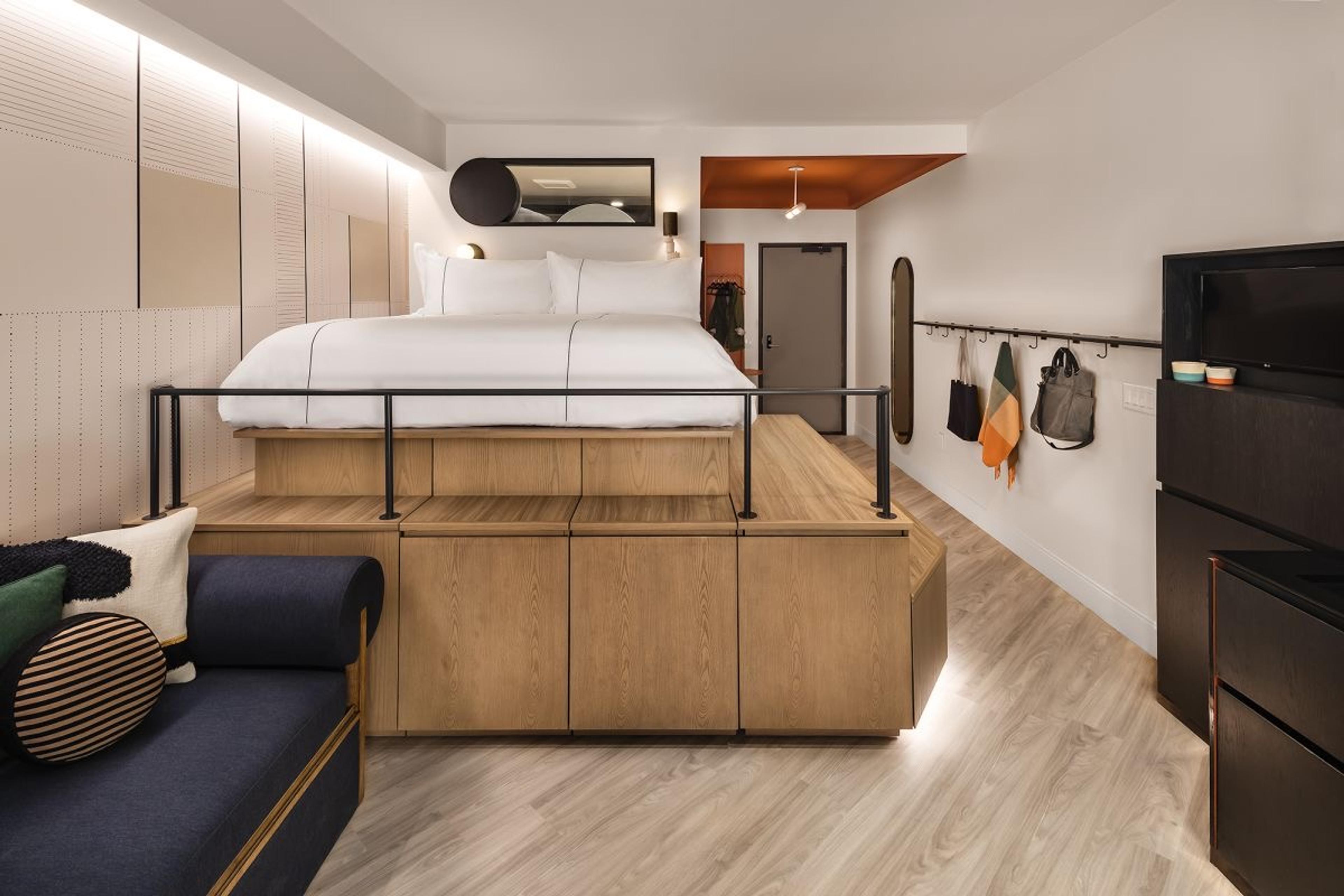 A hotel room with a bed set on a wooden platform above built-in drawers, a navy sofa in the foreground and a black cabinet against the far wall.