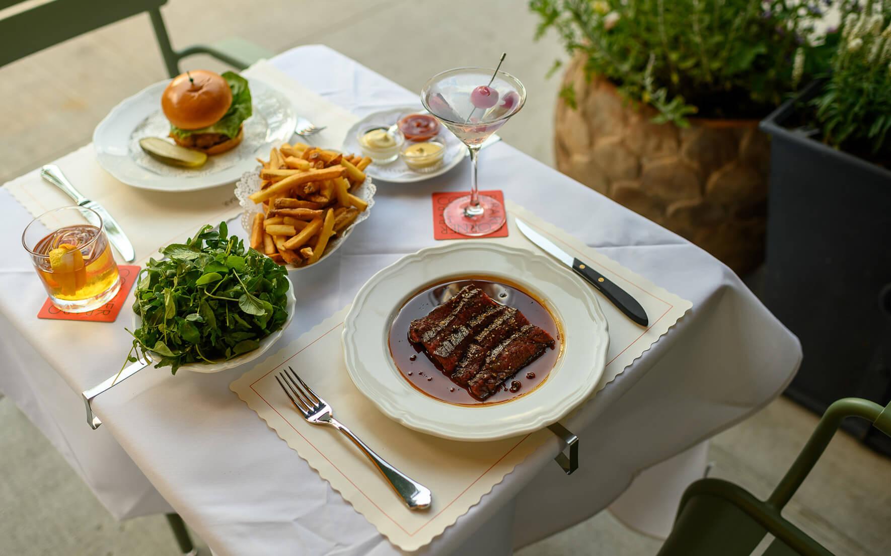 A table set with a burger, a bowl of fries, a bowl of greens, a plate of ribs, two cocktails and a glass of iced tea, with potted plants beside it.