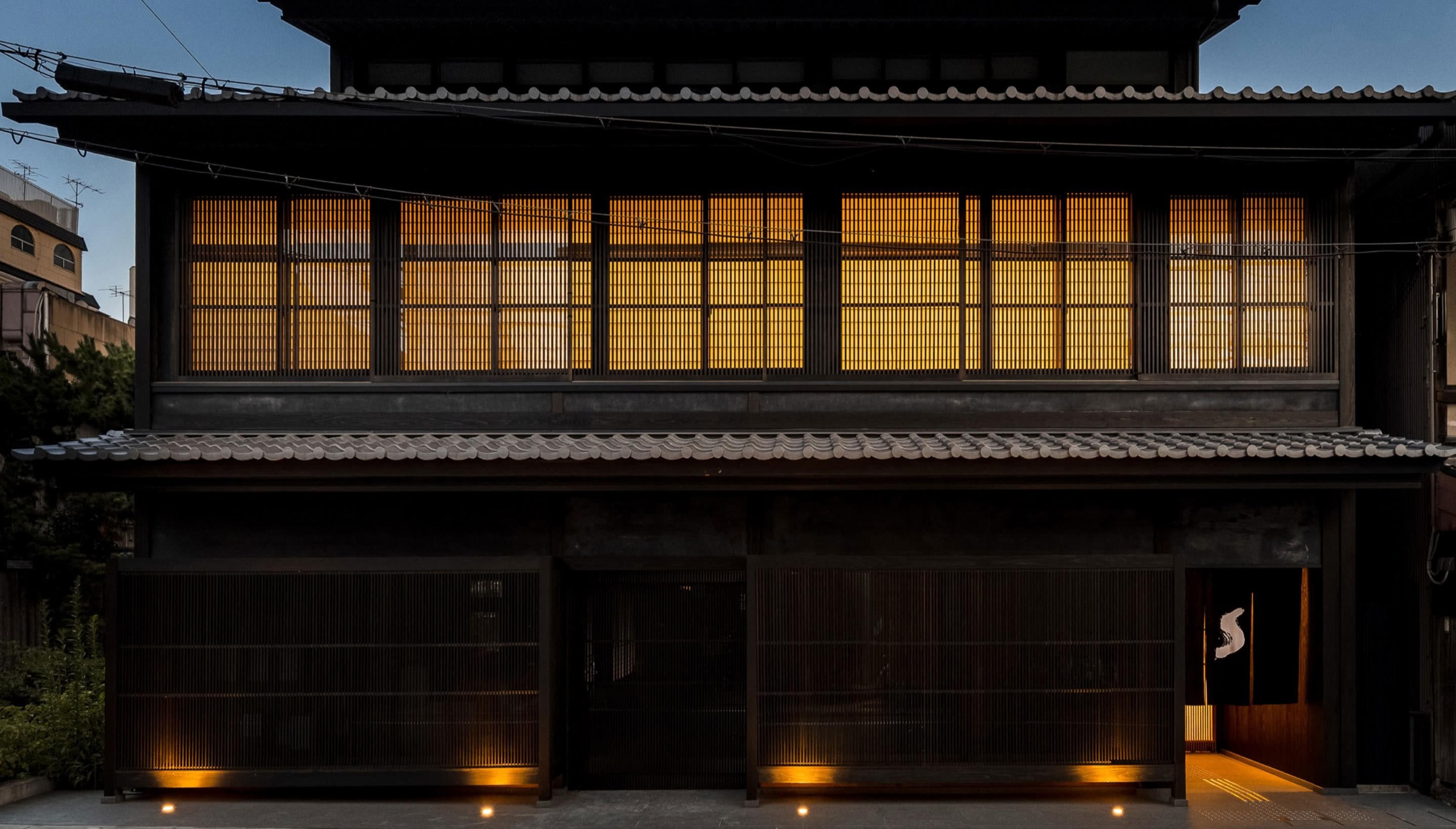 a two-storey Japanese timber townhouse at dusk, its upper windows glowing amber behind paper screens, with a lit entrance and an indigo curtain on the right