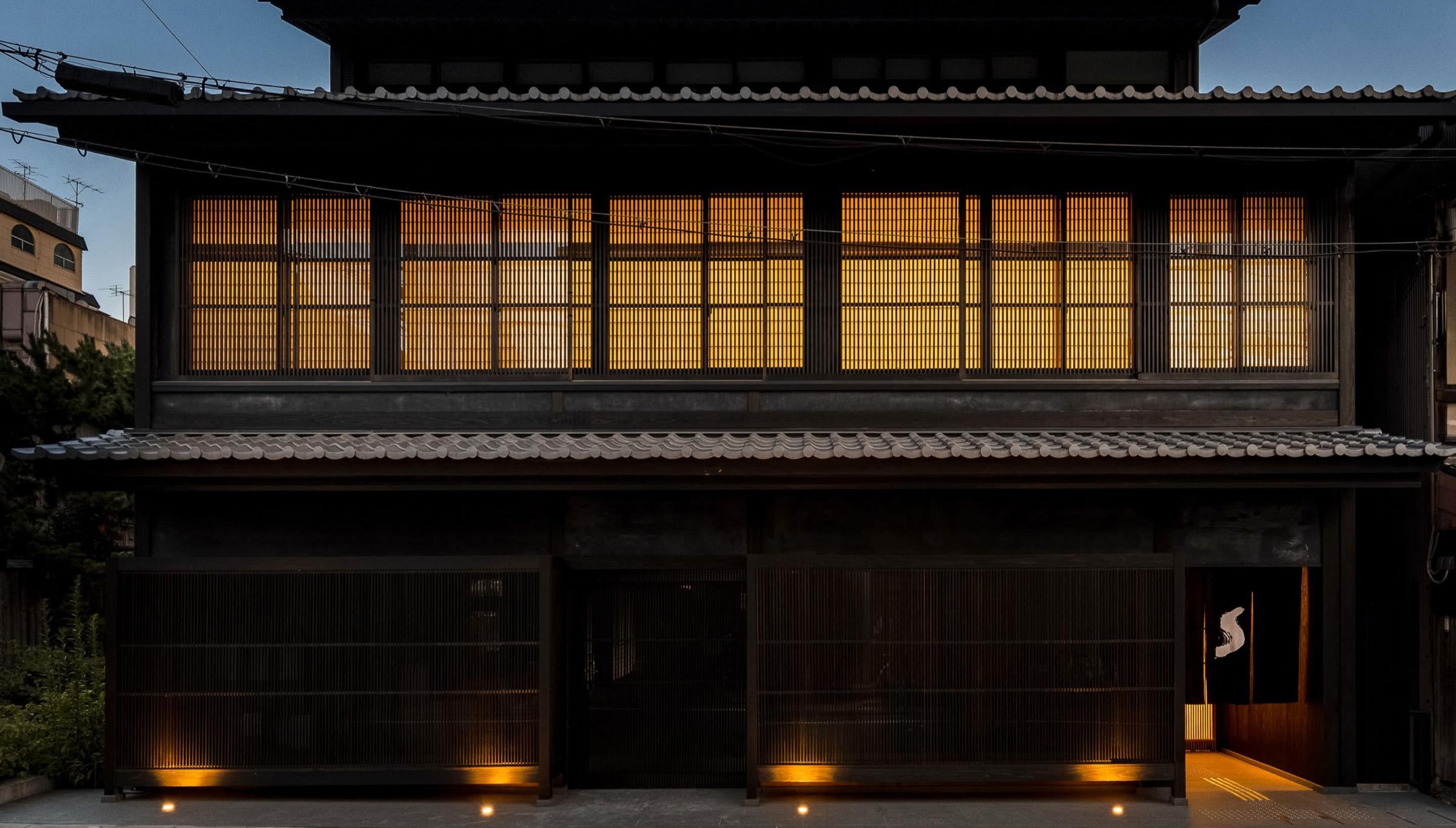 a two-storey Japanese timber townhouse at dusk, its upper windows glowing amber behind paper screens, with a lit entrance and an indigo curtain on the right