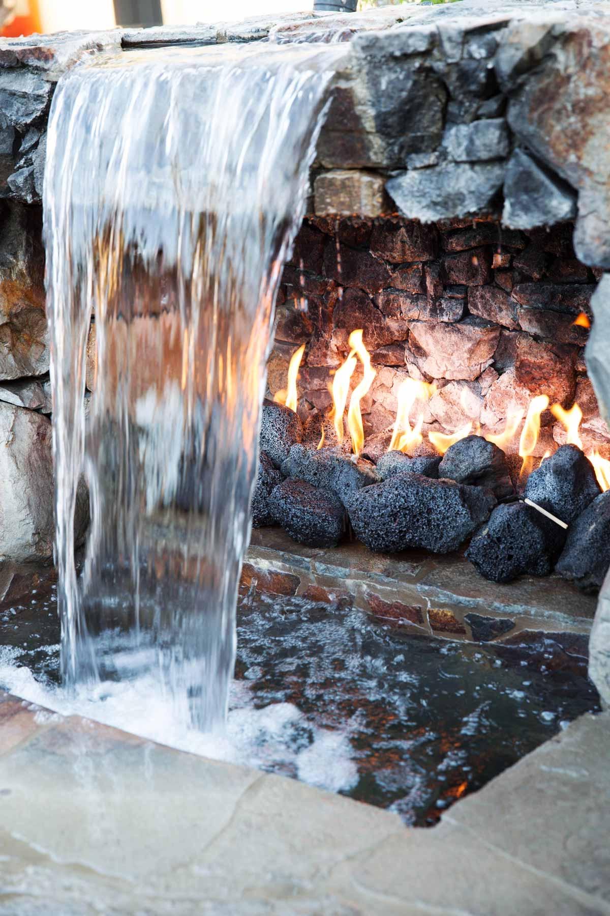 Water falls from a stone spout beside a row of gas flames in a stone recess, pooling in a flagstone basin below.