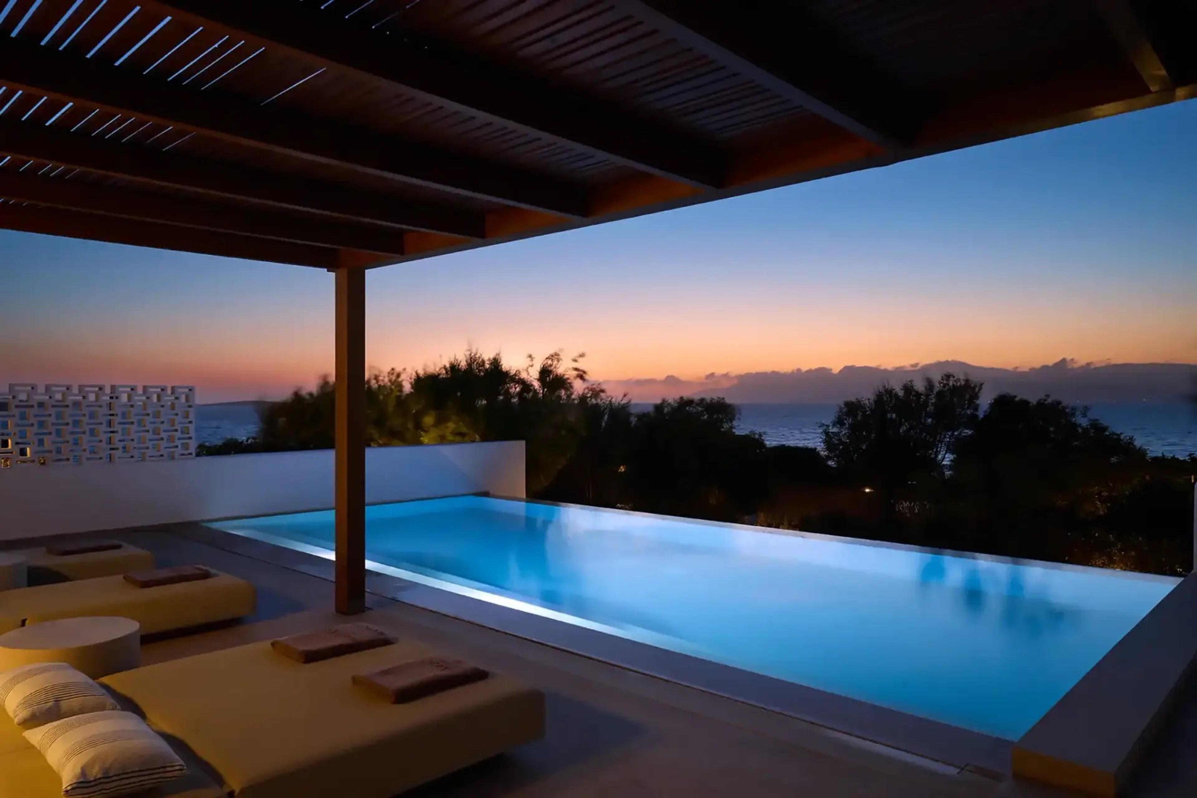 A pergola-covered terrace with daybeds beside a lit pool at dusk, the sky orange over the sea.
