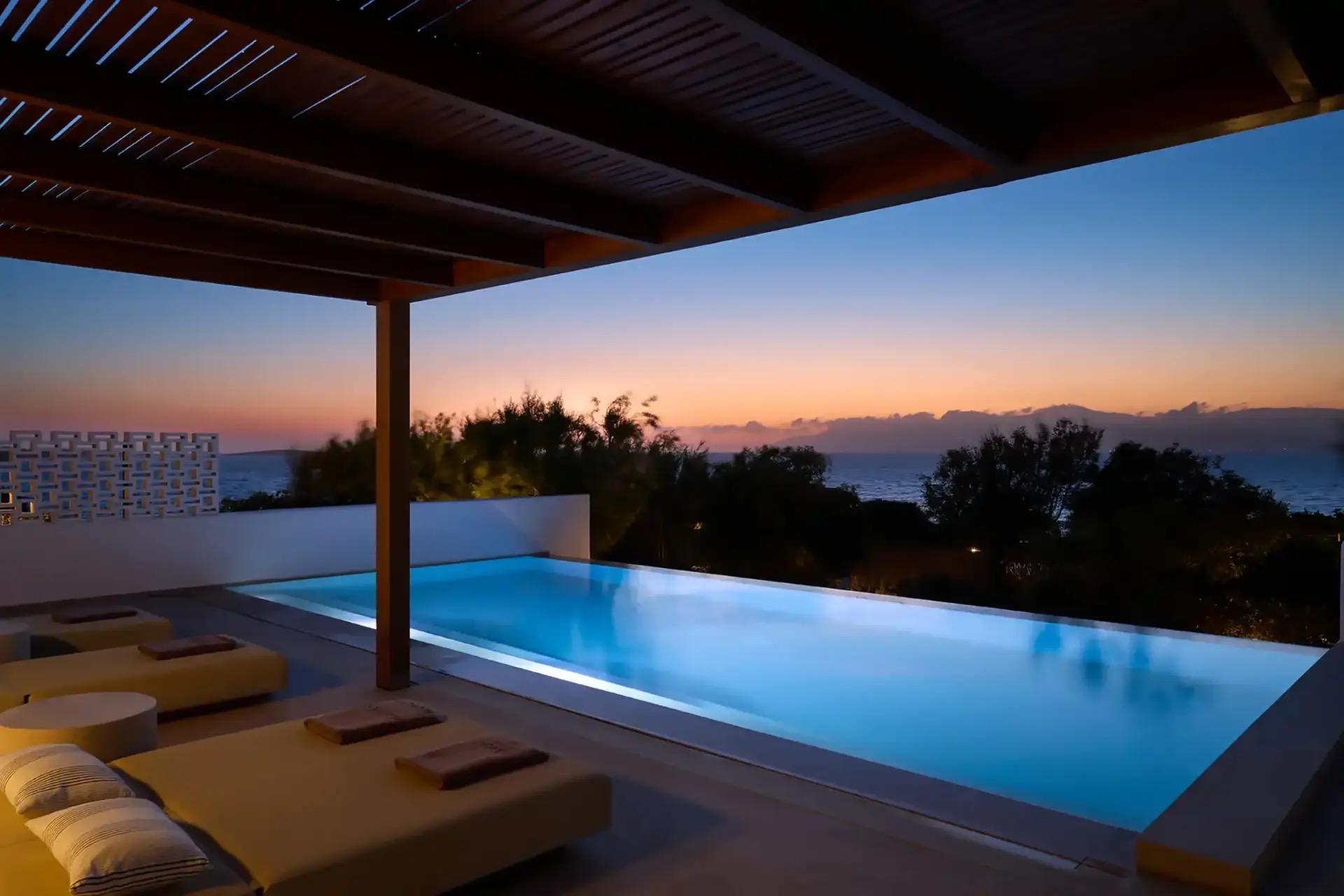 A pergola-covered terrace with daybeds beside a lit pool at dusk, the sky orange over the sea.