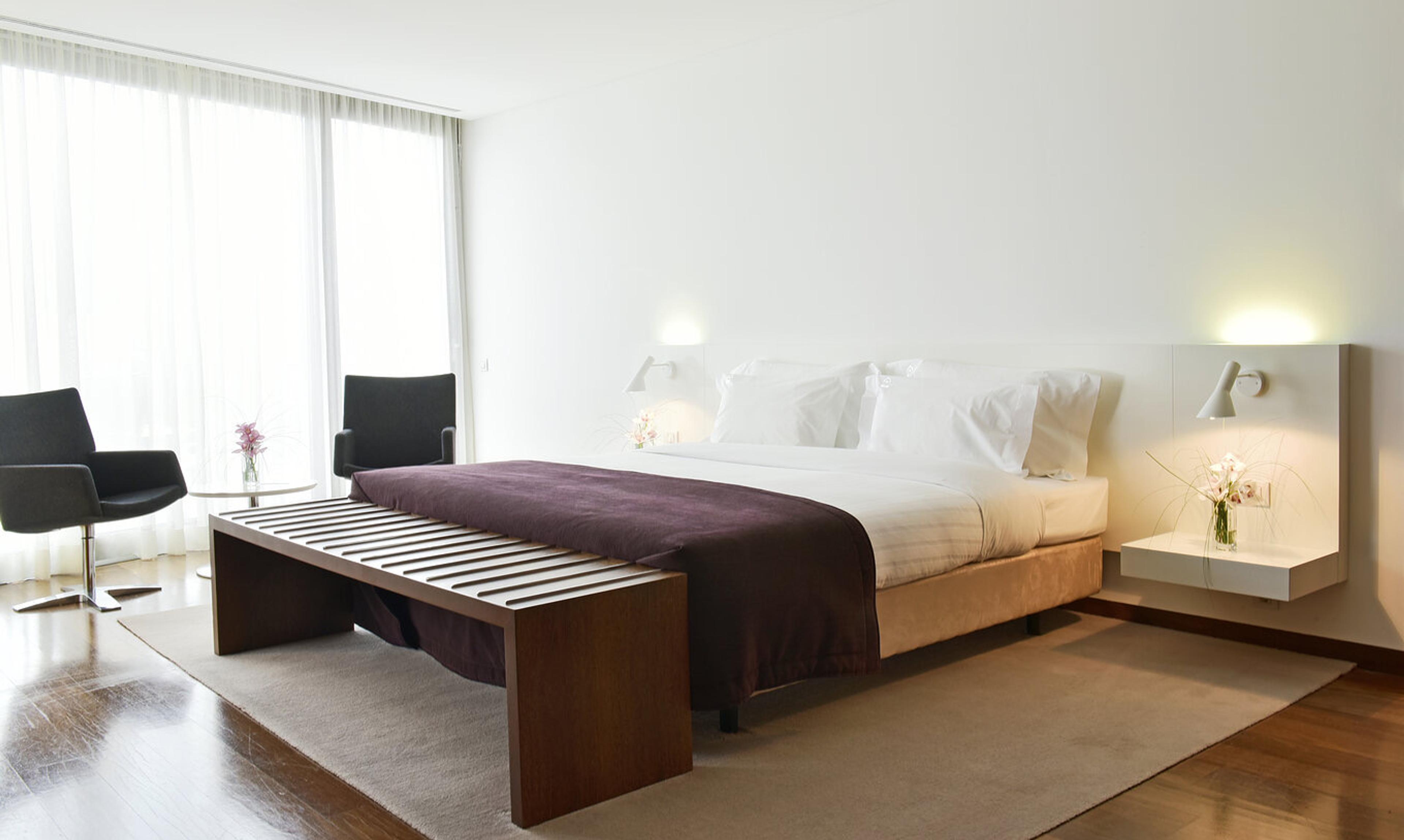 A bedroom with a double bed, a long wooden bench across the foot of it, and a window with sheer curtains and two armchairs.