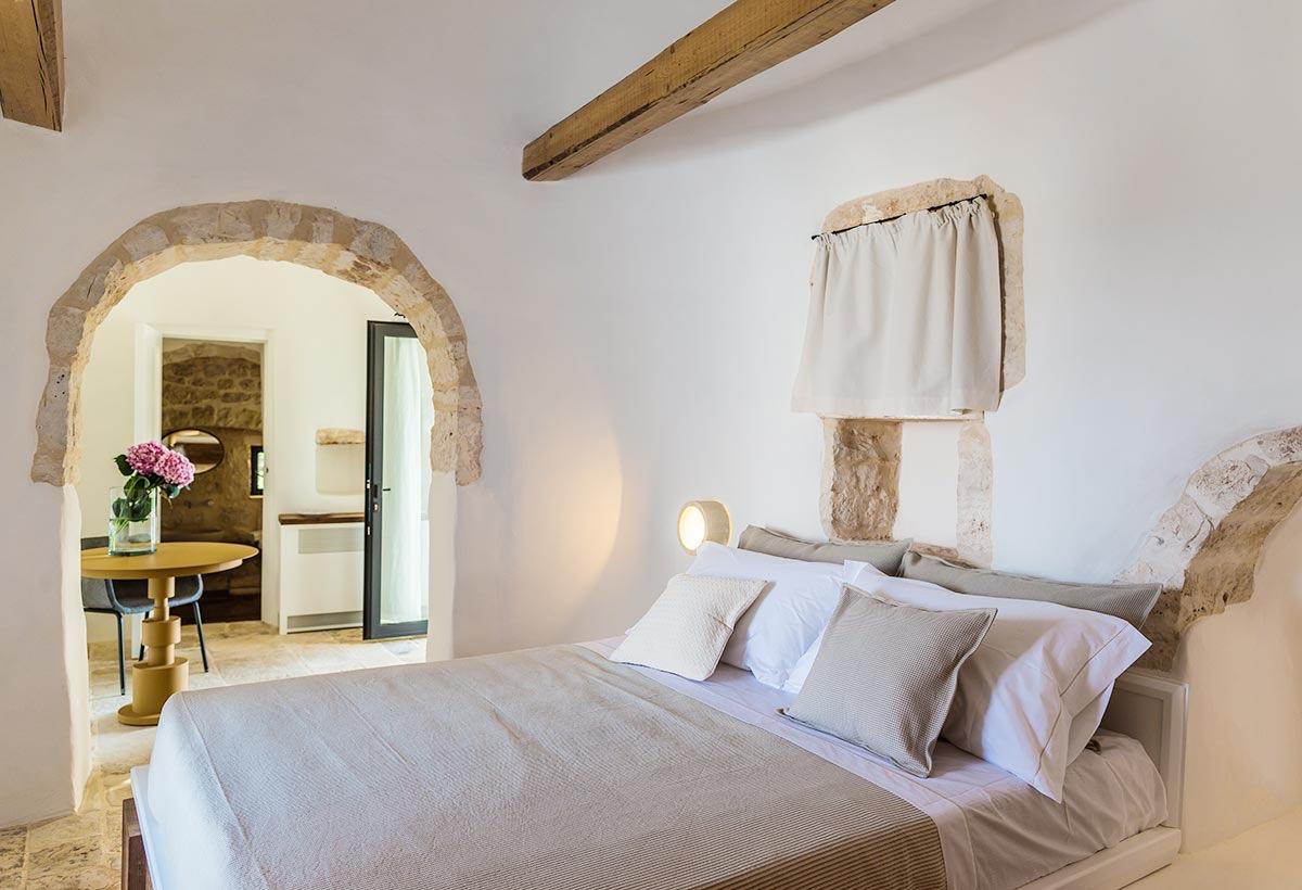 A bed with pale linen in a stone-arched room, with an open archway beyond showing a small table set with flowers.