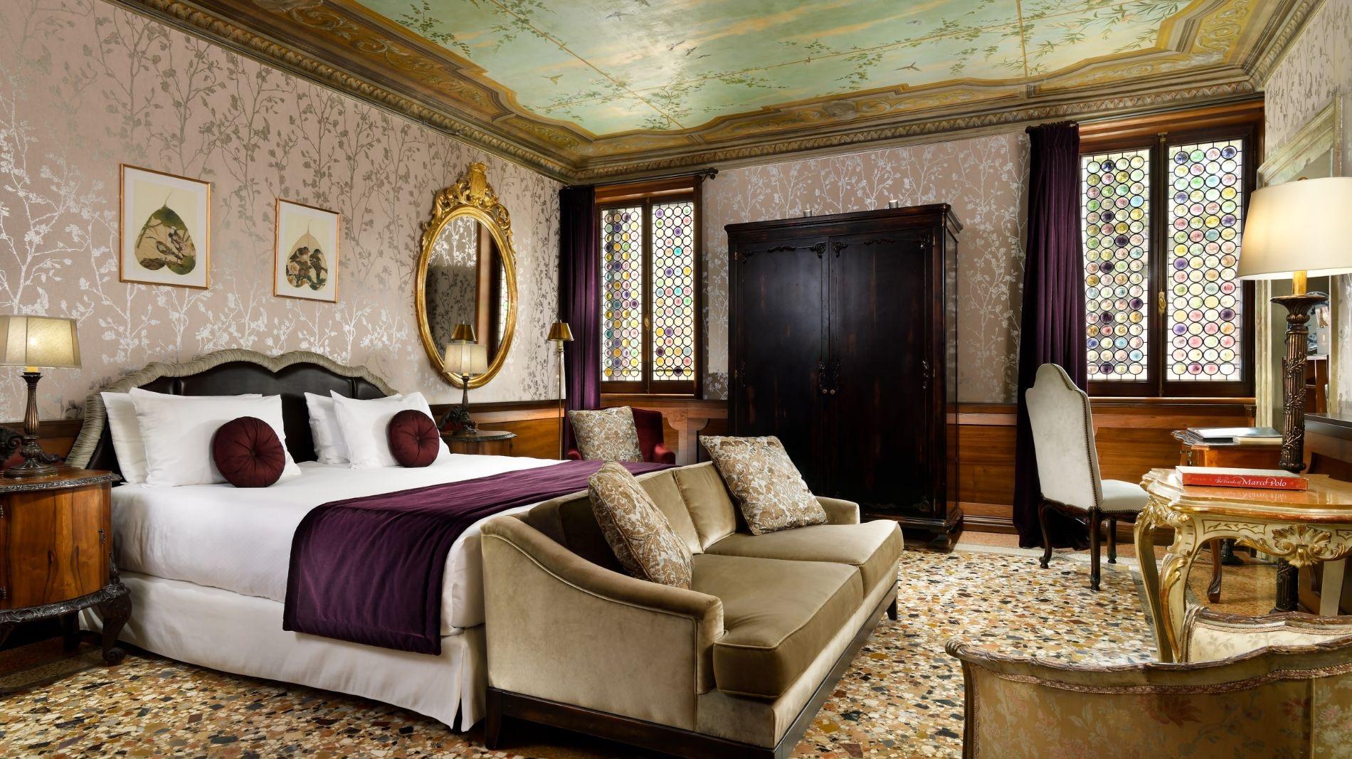 A bedroom with a painted and gilded ceiling, patterned wallpaper, a tufted headboard, a dark wood armoire and a beige sofa.