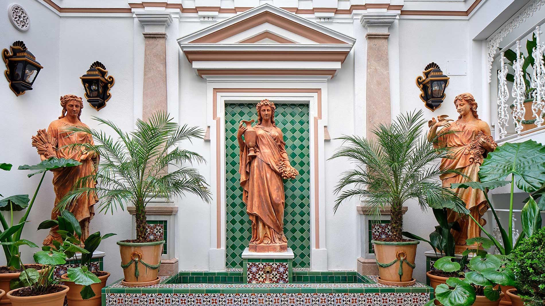 Terracotta statues of two women flank a fountain and potted palms in a white courtyard with green tiled alcoves.