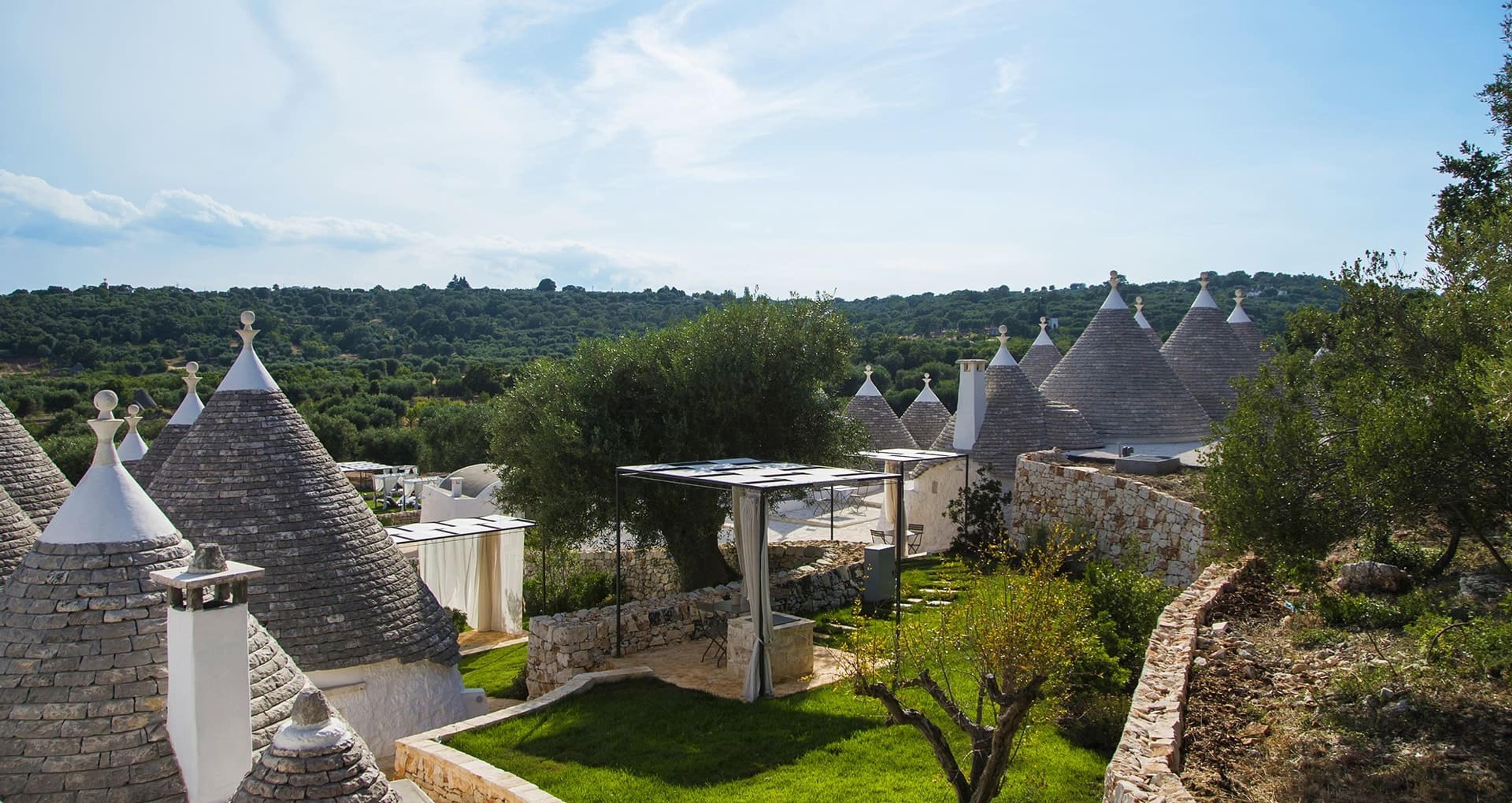 Stone cone roofs of several trulli rising above a walled garden and pergola, with an olive grove reaching to the horizon.