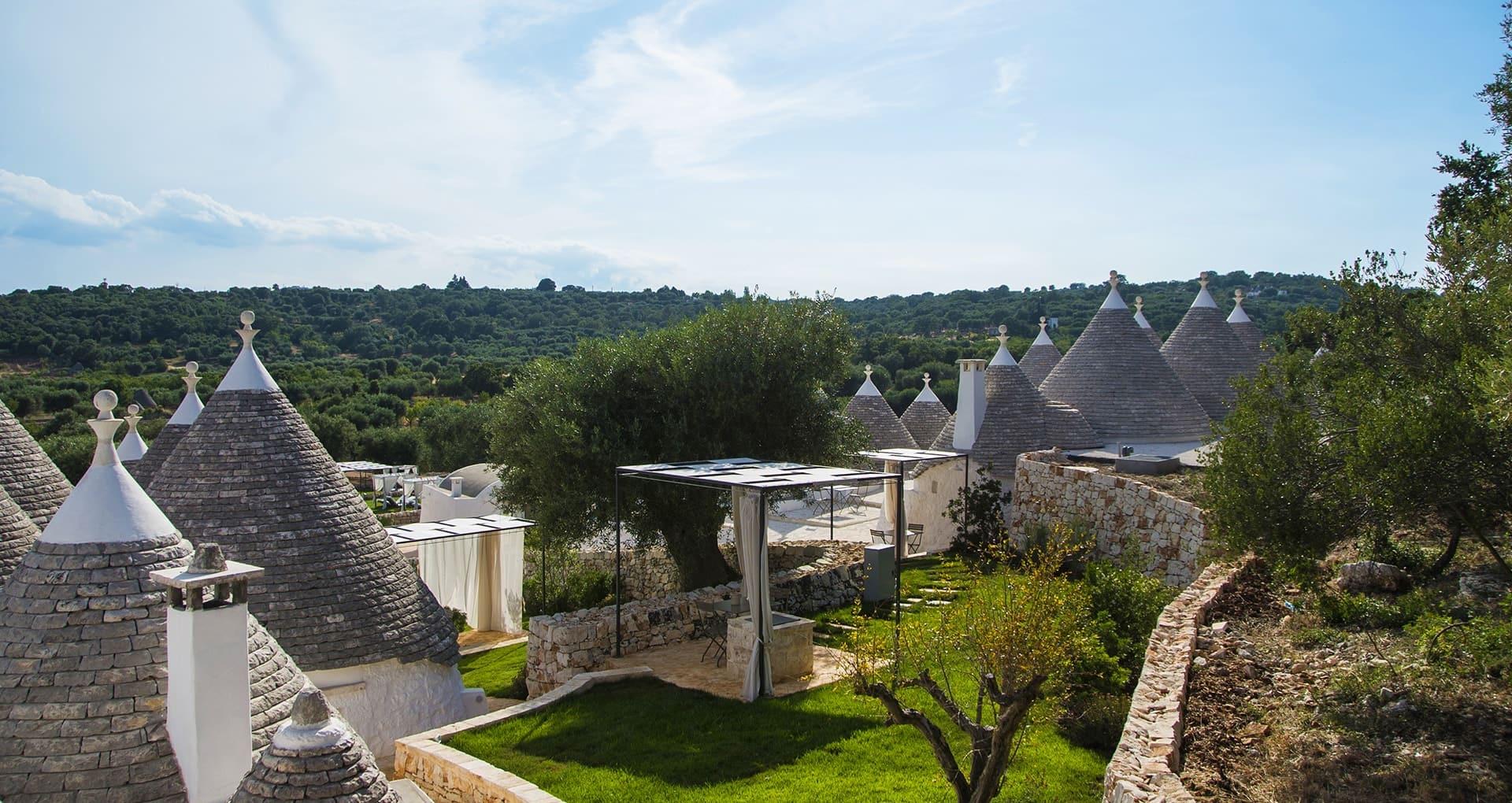 Stone cone roofs of several trulli rising above a walled garden and pergola, with an olive grove reaching to the horizon.