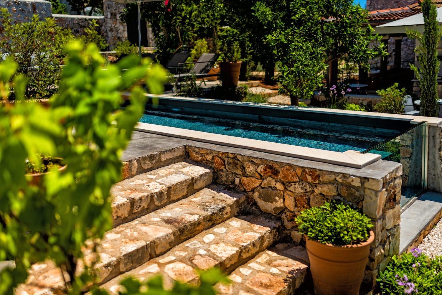 Stone steps leading down past a potted plant to a narrow pool with a pergola and the stone house behind.