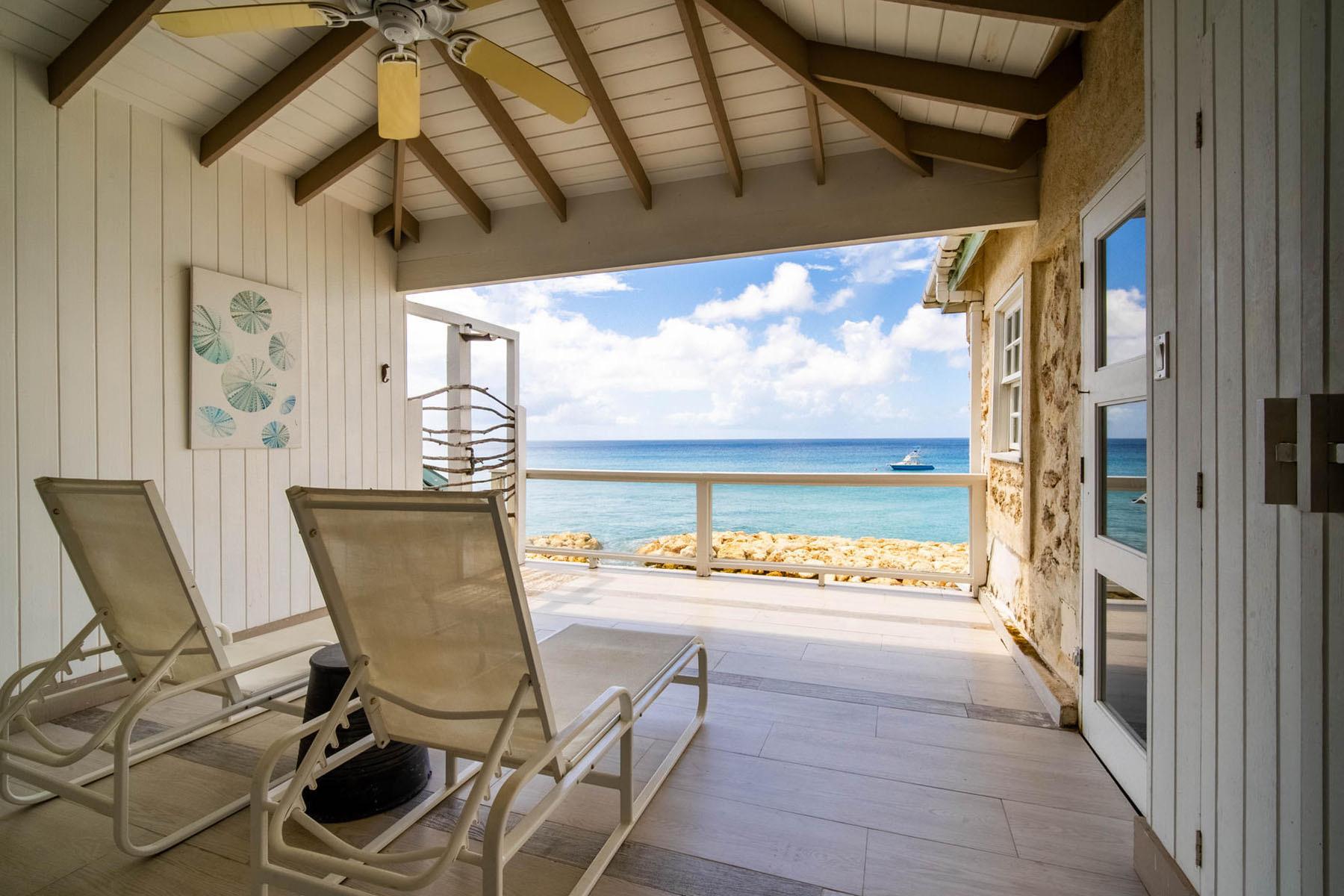 Two empty sun loungers on a covered stone terrace, facing a wide view of the sea over a low railing.