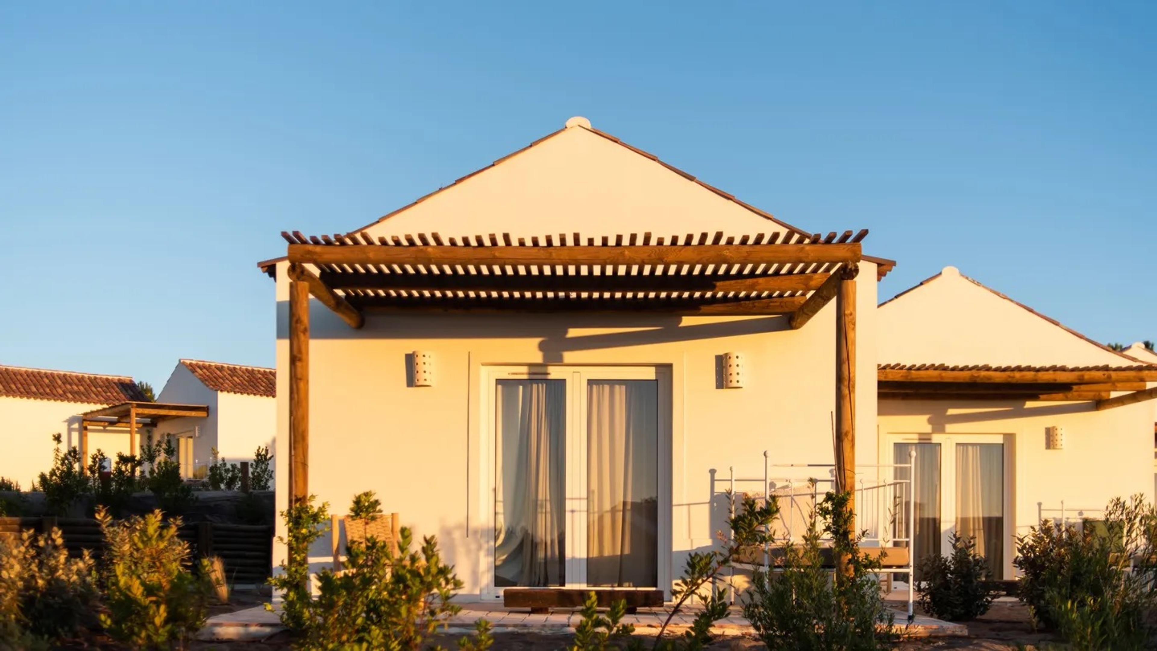 a white villa with a pitched tile roof and a wooden pergola shading its terrace, glass doors and curtains lit at dusk