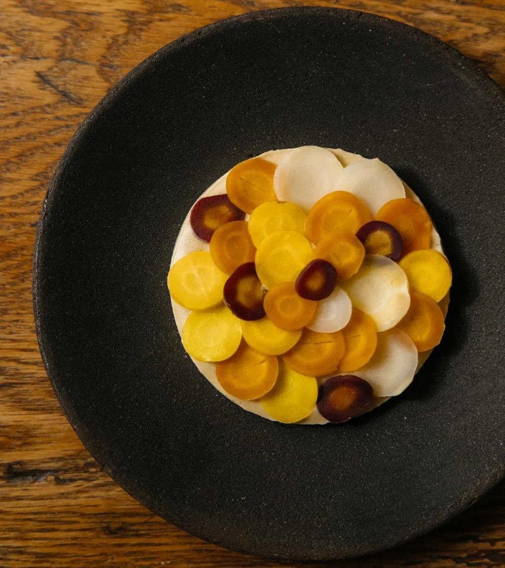 Rounds of yellow, orange, purple and white carrot laid over a cream base on a dark stoneware plate at Rosetta