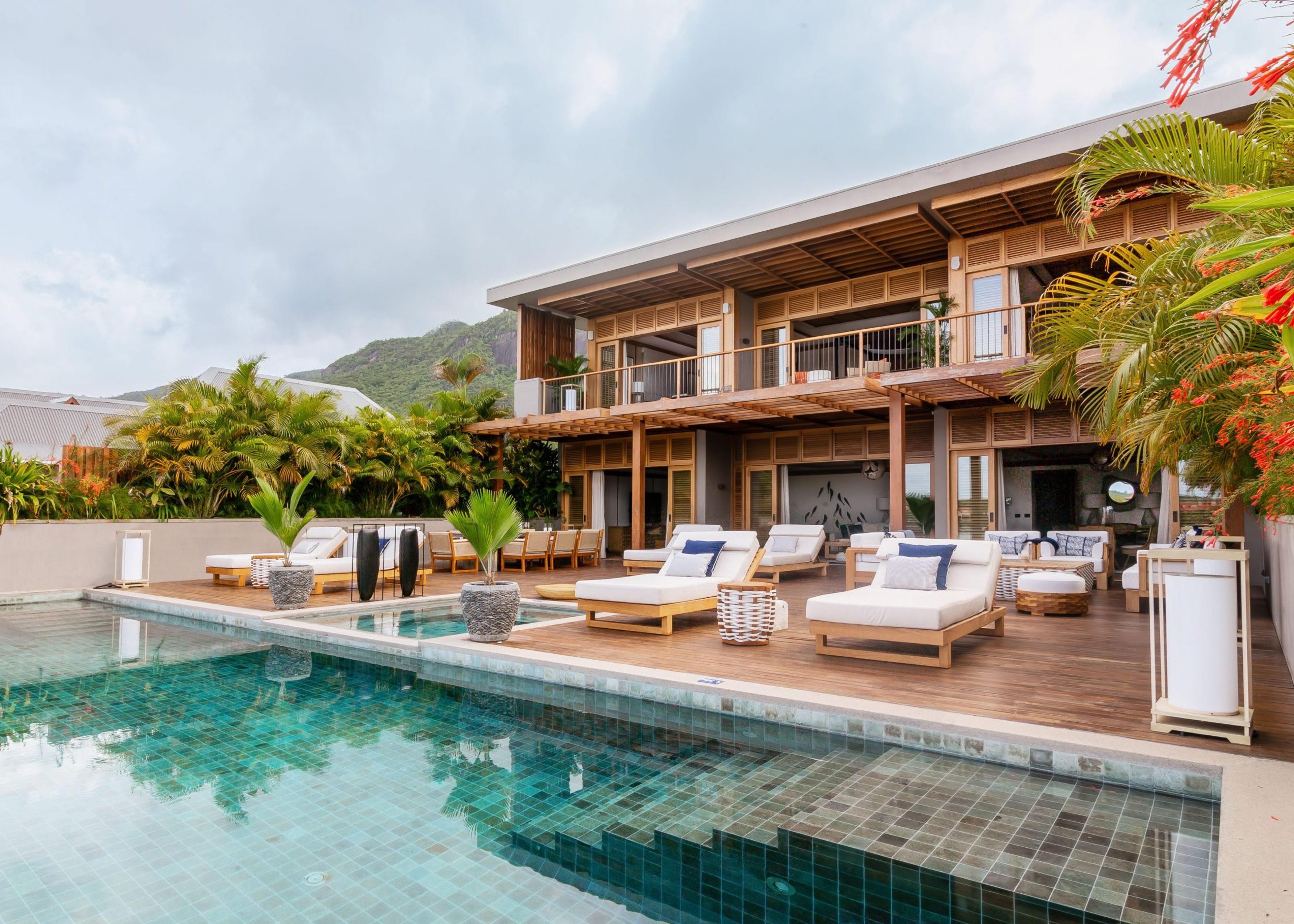 A two-story wooden hotel building behind an infinity-edge pool, with forested hills in the background and sun loungers along the deck.