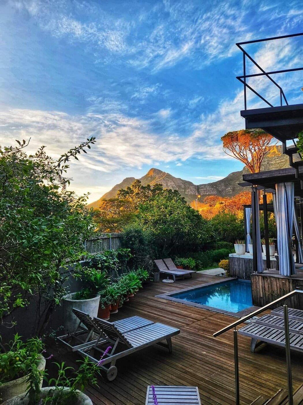 A timber deck at sunset beside a small blue plunge pool, with two loungers, potted plants, a steel-railed balcony above, and a mountain ridge behind the trees.