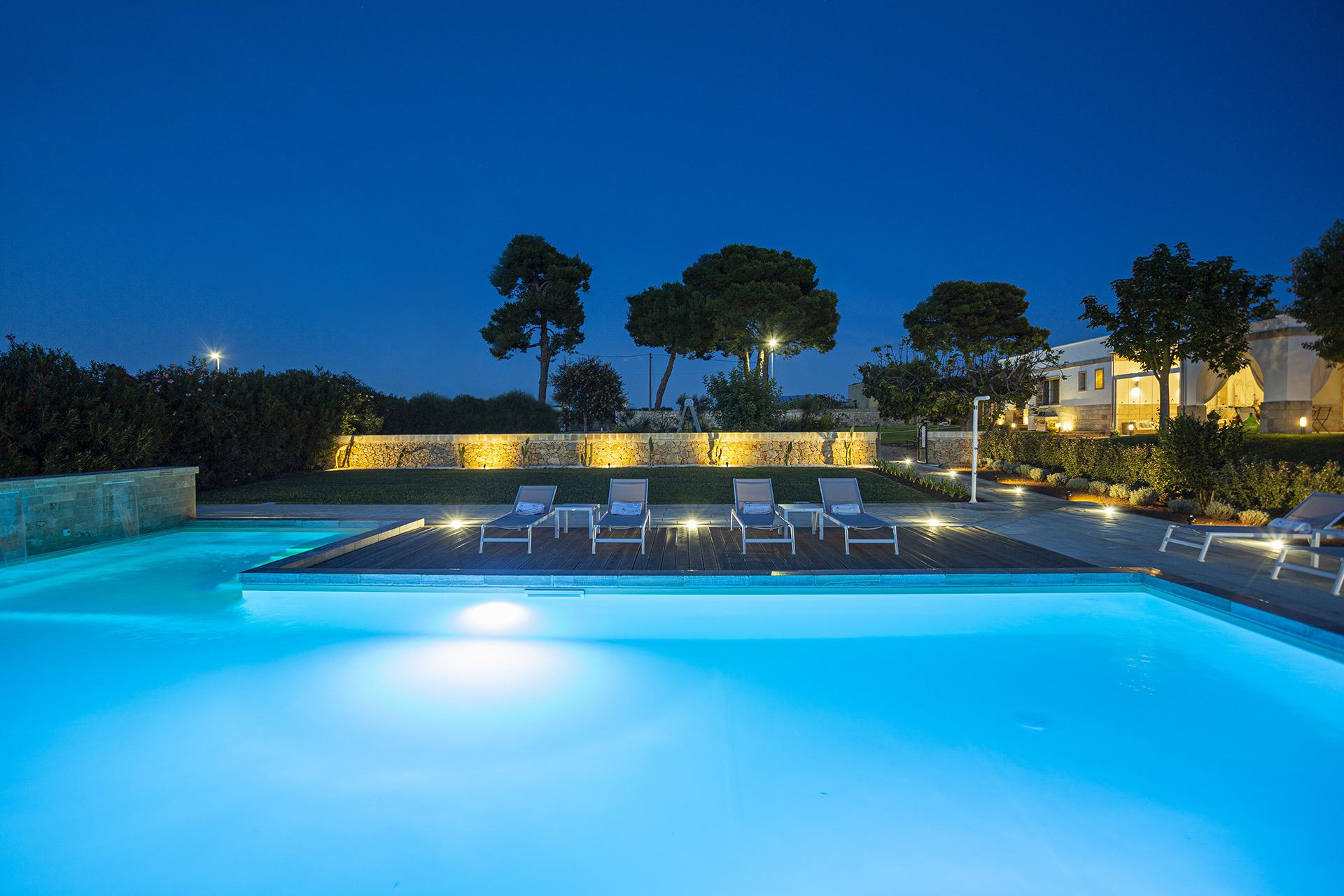 A pool lit blue at dusk in front of the stone-walled hotel building, its windows lit and loungers set out on a wooden deck.