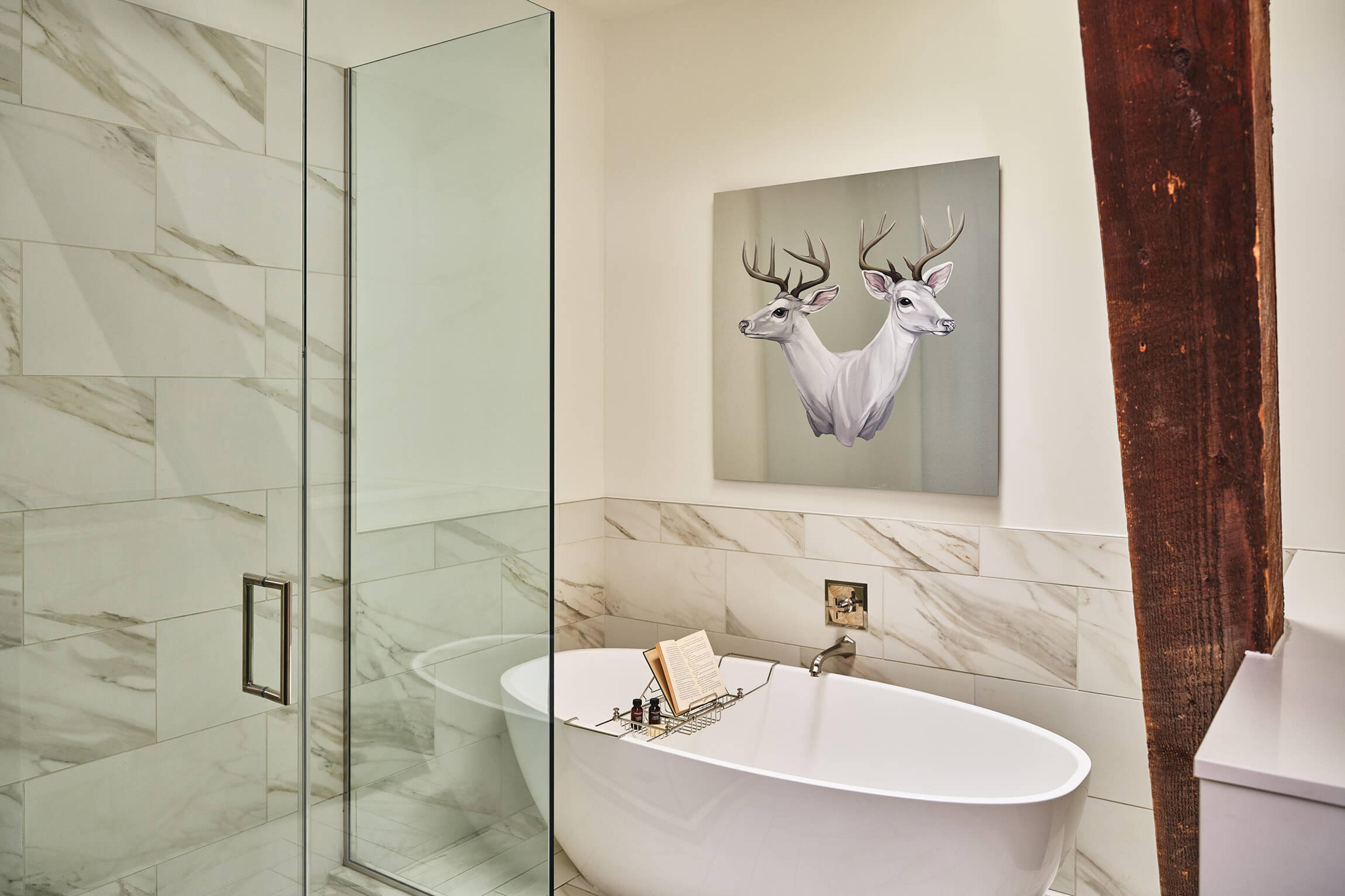 A glass-walled shower and a freestanding white bath on marble tiles, with a painting of two white deer above the bath and a wooden beam on the right.