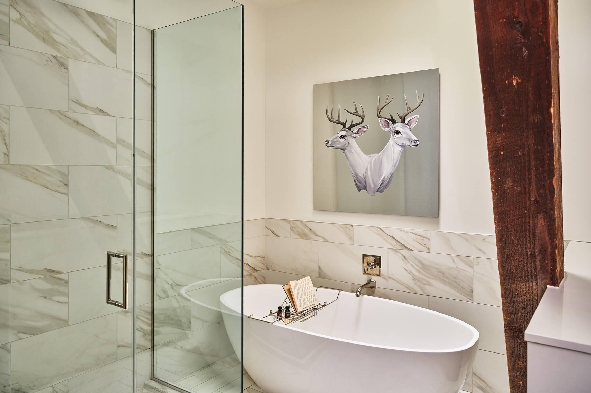 A glass-walled shower and a freestanding white bath on marble tiles, with a painting of two white deer above the bath and a wooden beam on the right.