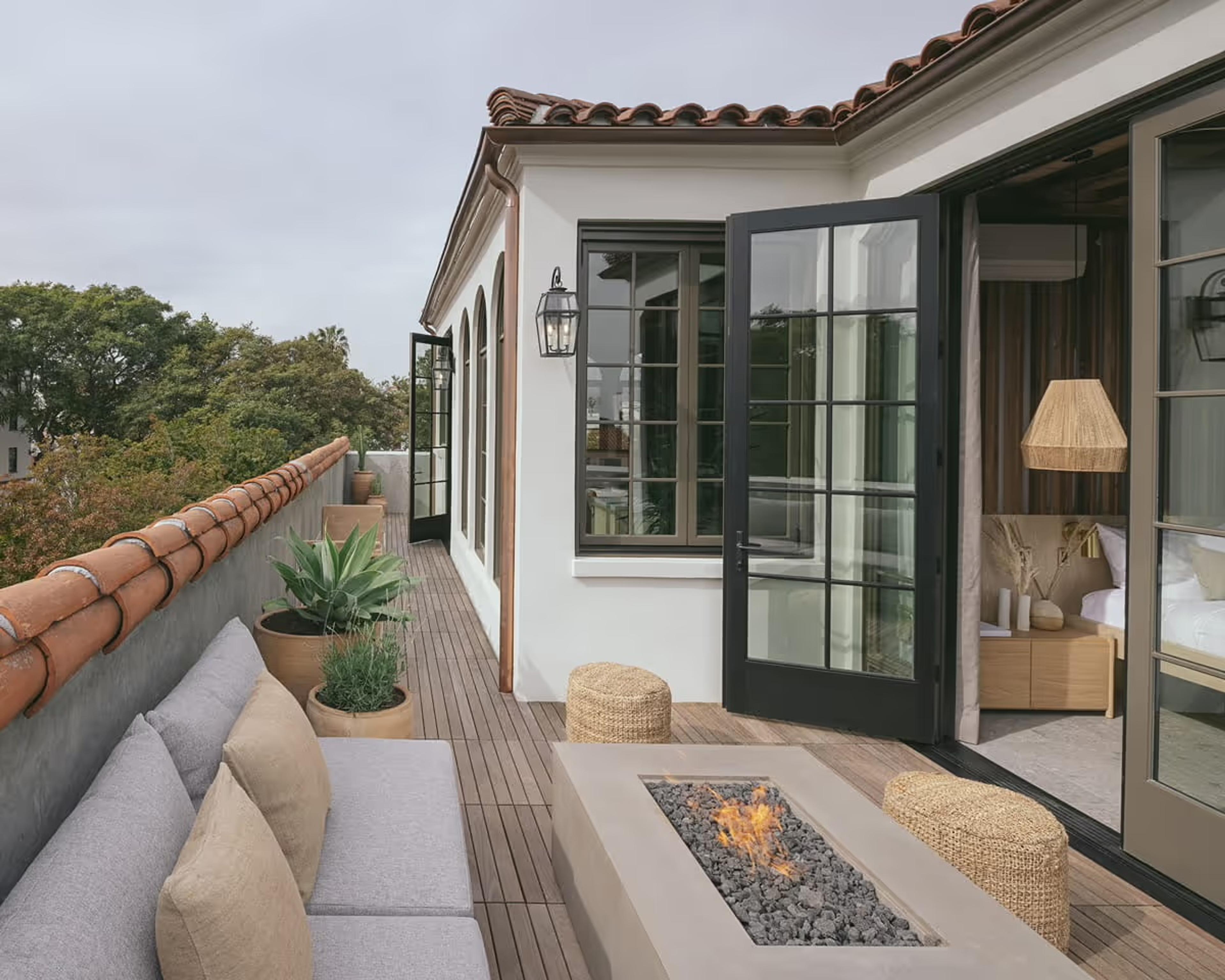 A rooftop terrace with a built-in grey sectional sofa, a lit stone fire pit, potted agave plants, and open French doors leading into a bedroom under a Spanish tile roof.
