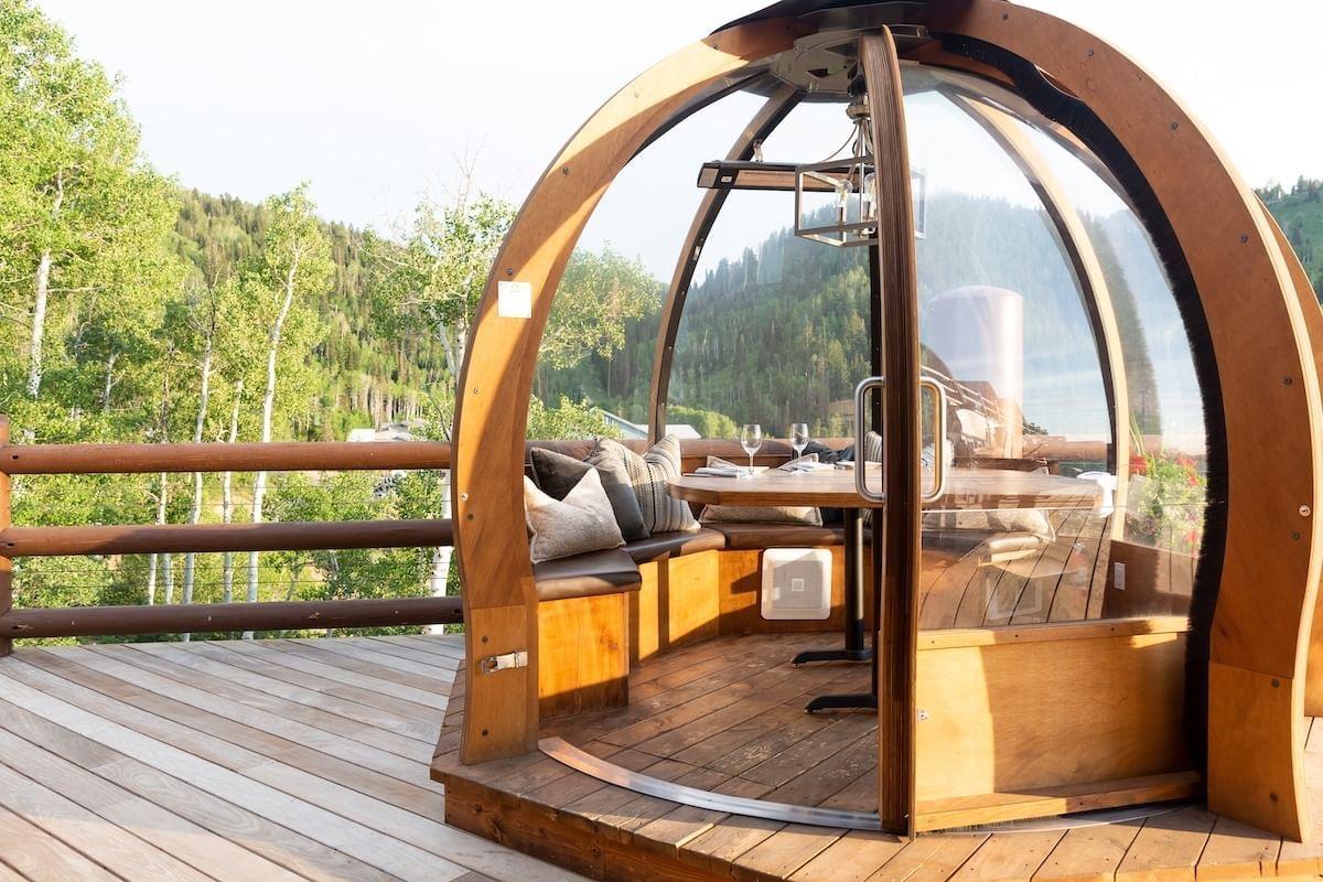 a wood framed glass dome on a deck, with a table set for two laid with wine glasses, looking out over aspen trees