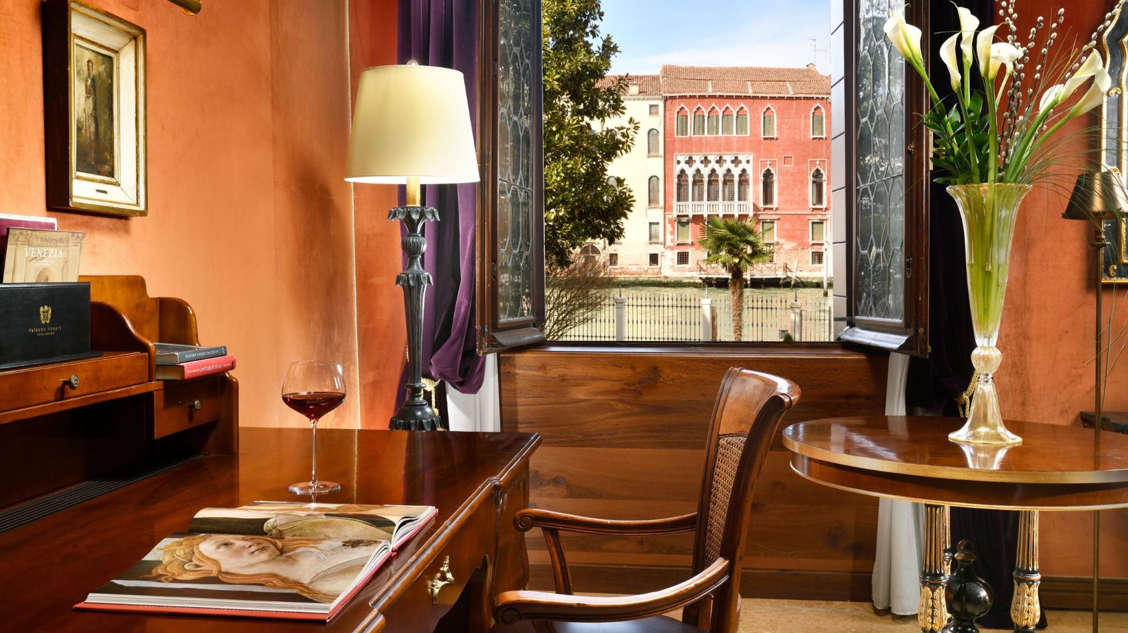 A writing desk by an open window with a glass of red wine, an open book and a vase of lilies, looking out over a canal to a red Venetian palazzo across the water.