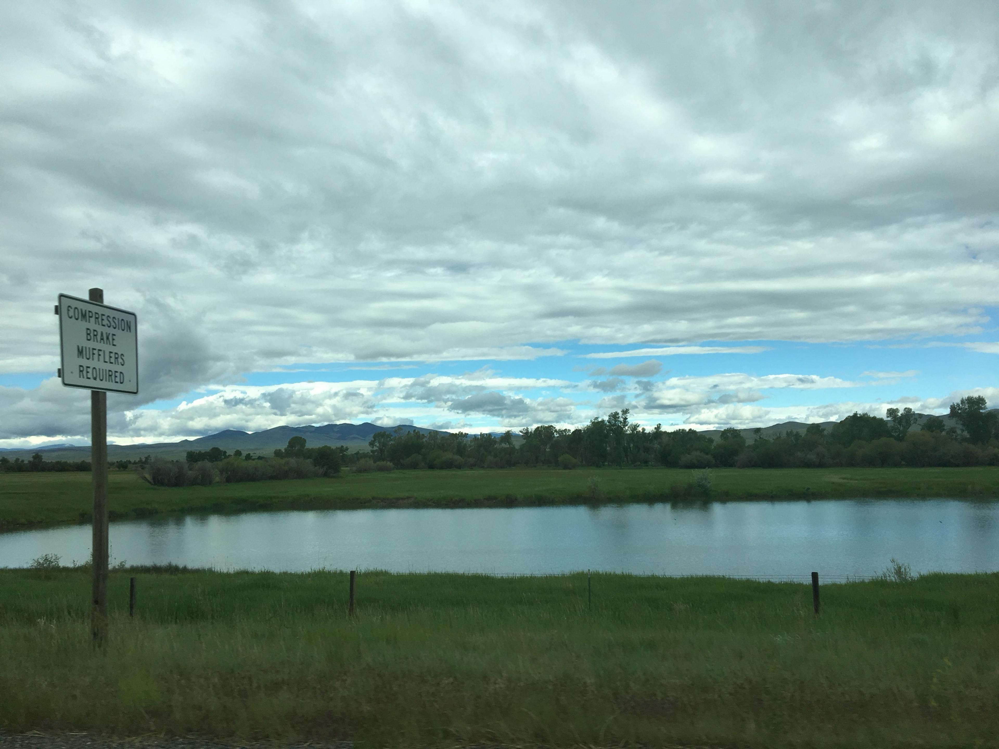 A pond in flat green ranchland under a heavy grey sky, with low mountains along the horizon