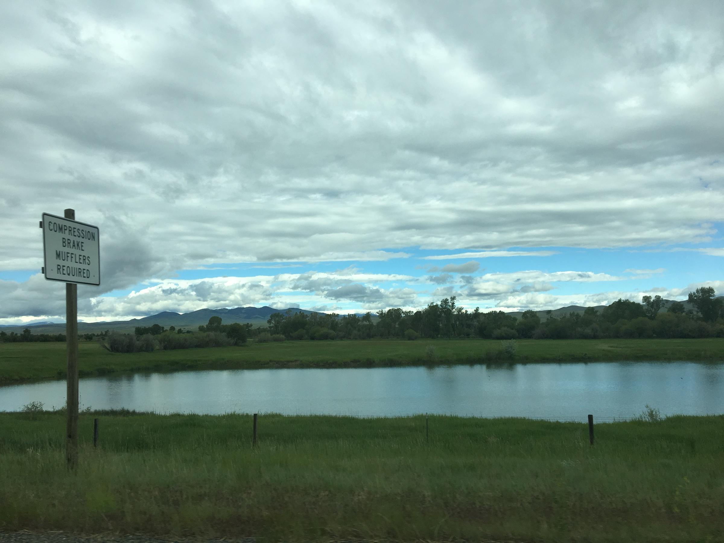 A pond in flat green ranchland under a heavy grey sky, with low mountains along the horizon