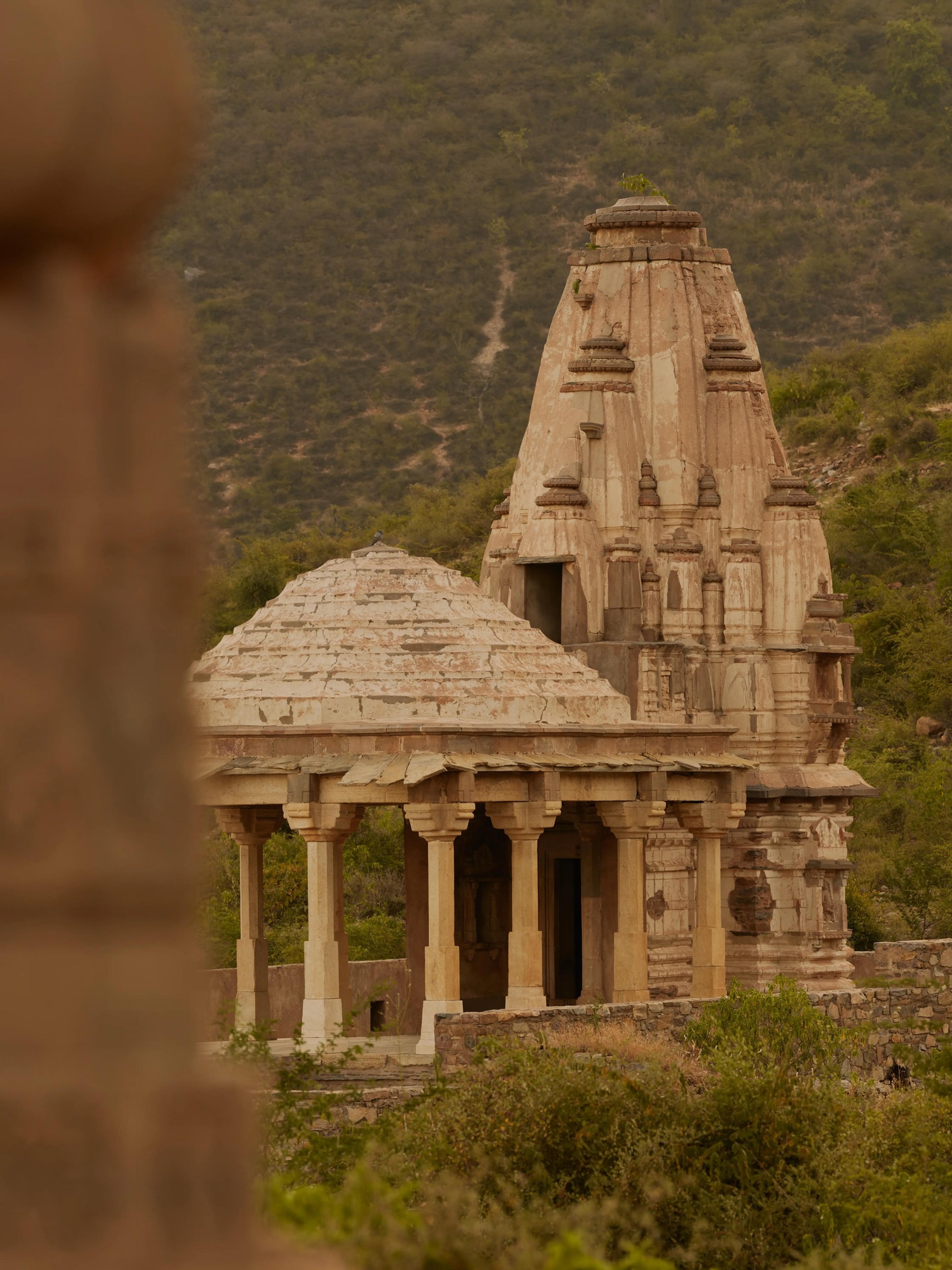 A carved stone temple with a tiered tower, seen past a domed pavilion on a hillside of scrub and trees.