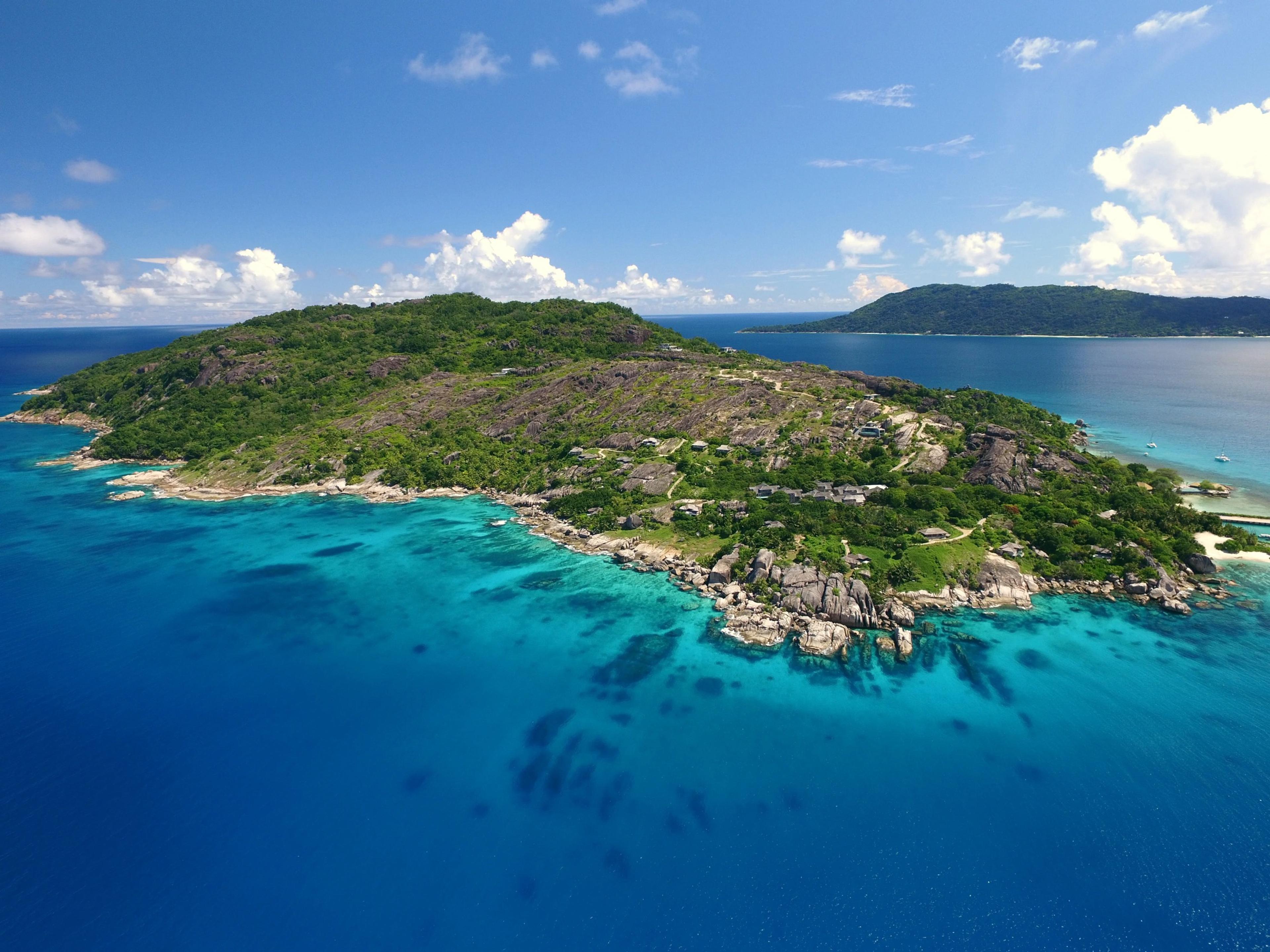 An aerial view of a green, rocky island surrounded by turquoise and deep blue ocean under a partly clouded sky.