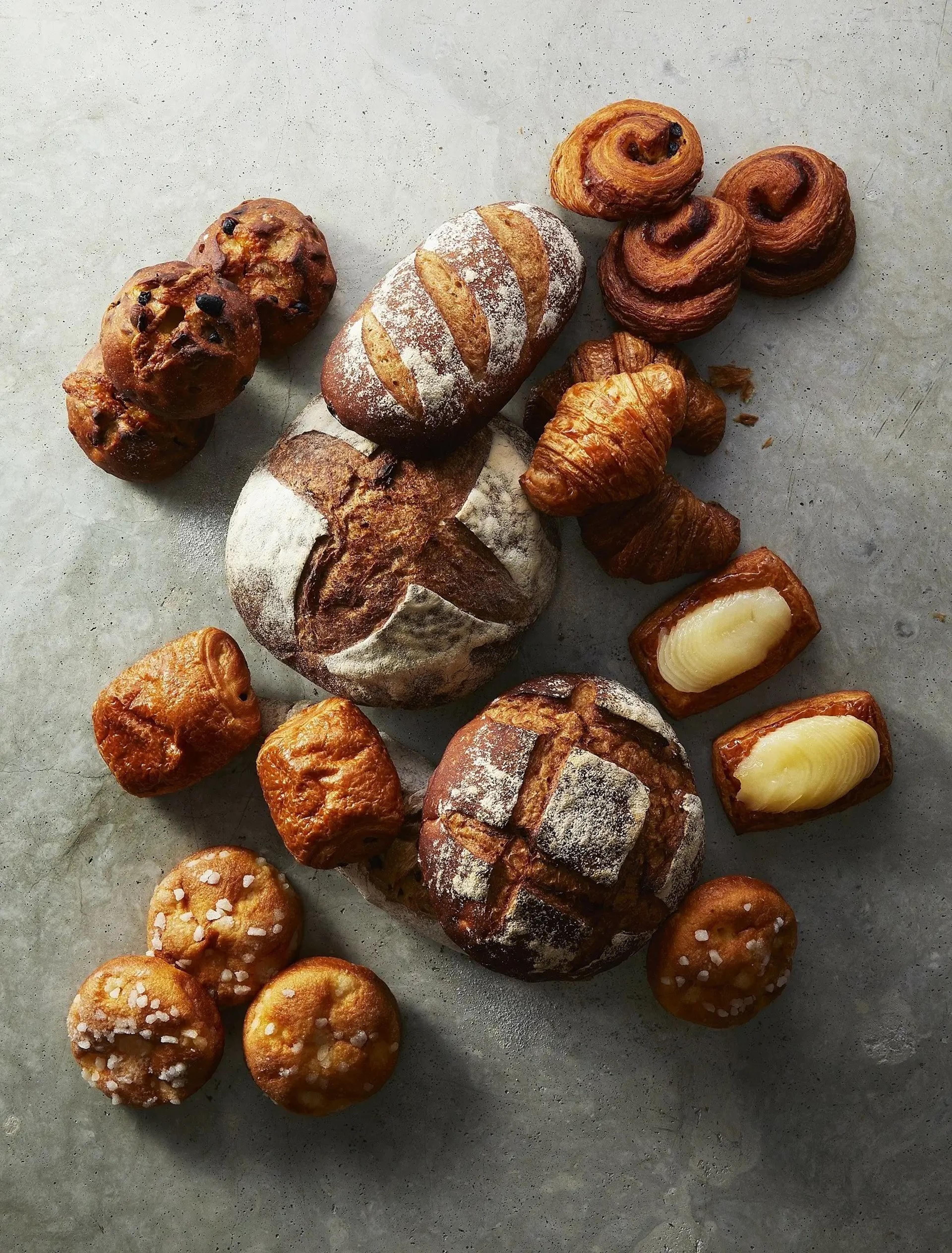 An overhead view of loaves, croissants, pastries and salted rolls laid out on a pale stone surface.