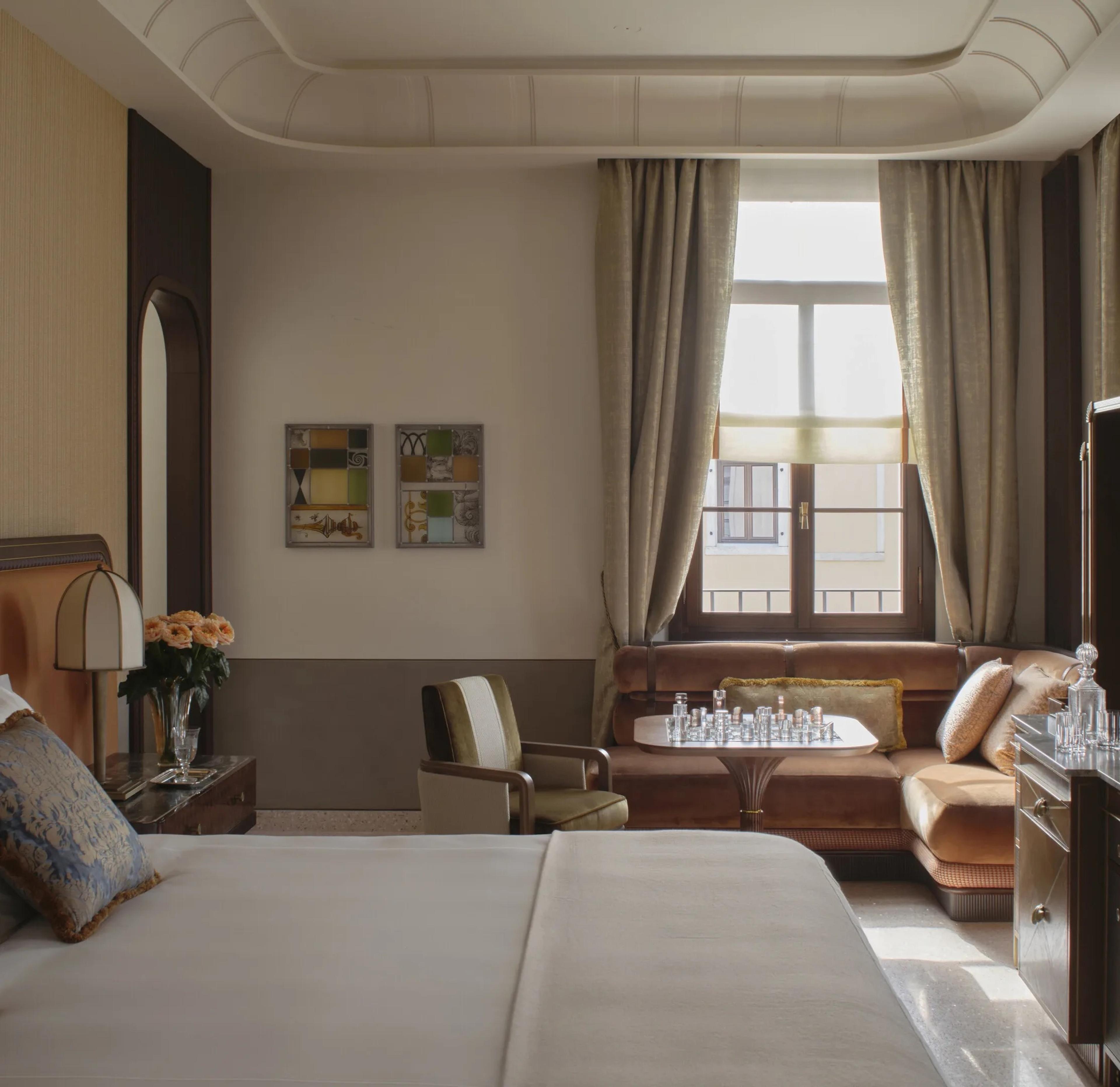 A hotel suite's sitting corner with a brown velvet sofa, an armchair, and a small games table set with a crystal chess set, beside a curtained window