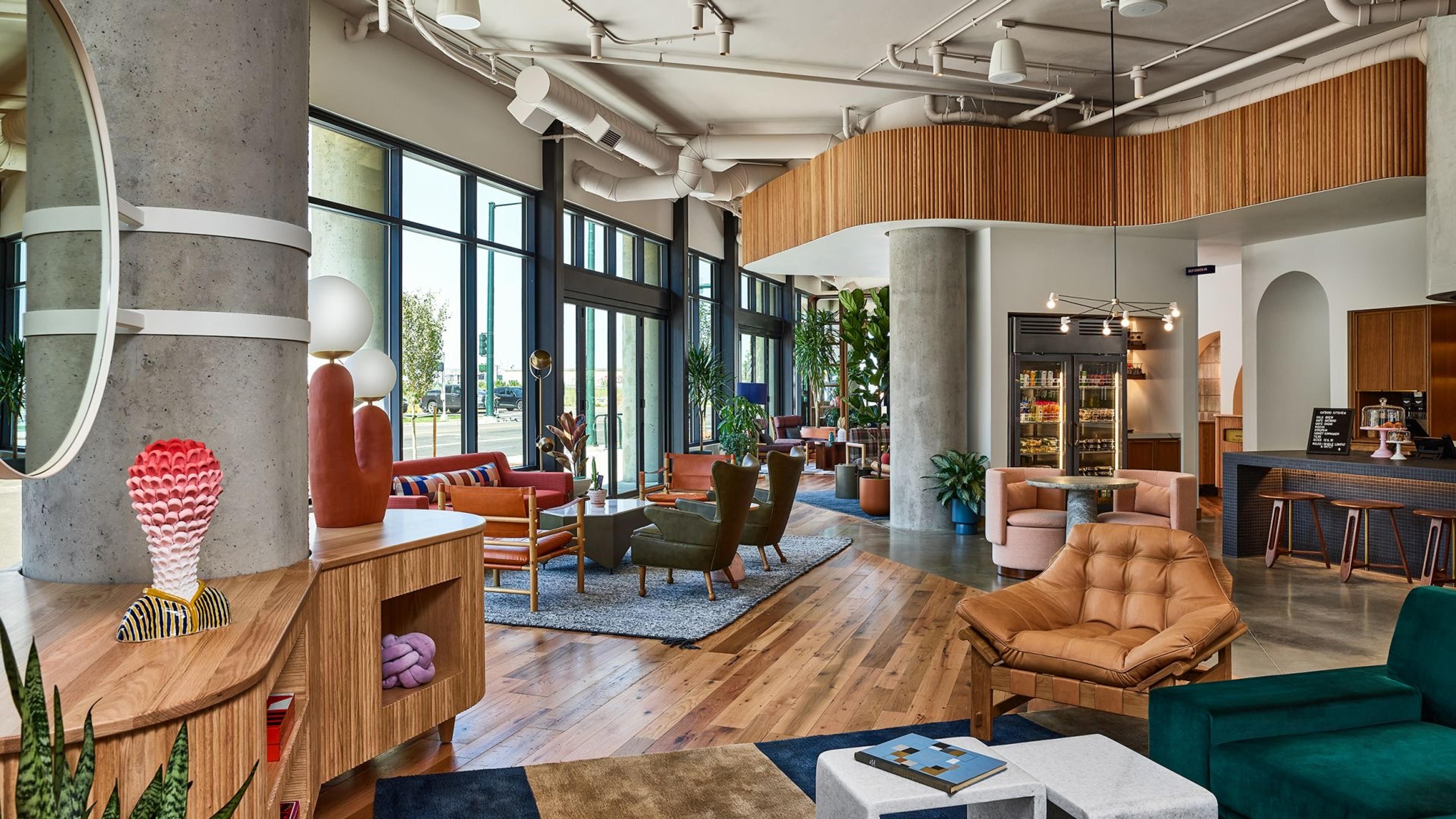 A hotel lobby and cafe with concrete columns, floor to ceiling windows, wood flooring and a mix of leather and velvet chairs around low tables.